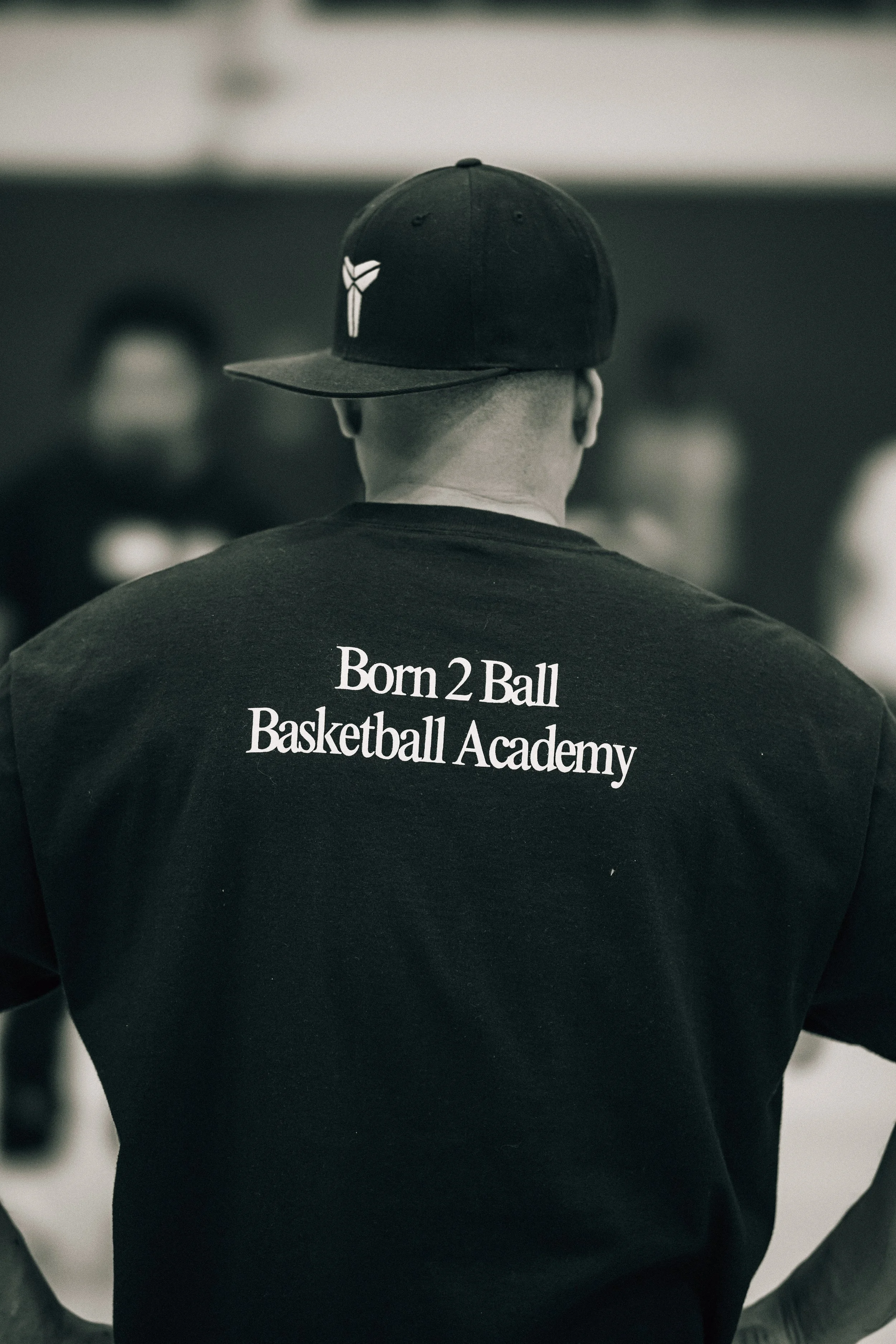 Back view of a person wearing a black cap and T-shirt with the text 'Born 2 Ball Basketball Academy' printed on the back, in a gym or sports facility setting.