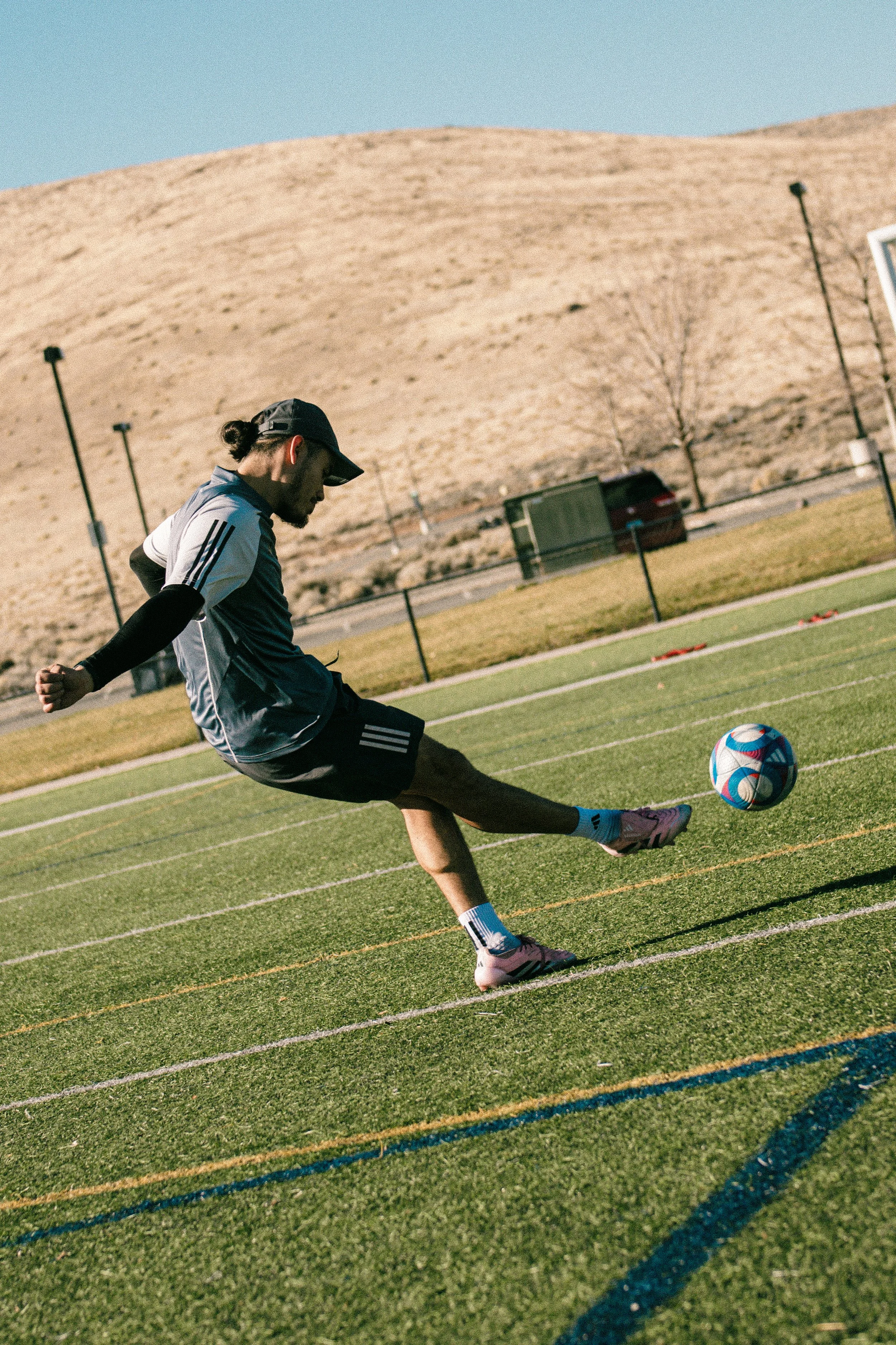 A person in athletic clothing kicking a soccer ball on a grassy field with hills in the background.