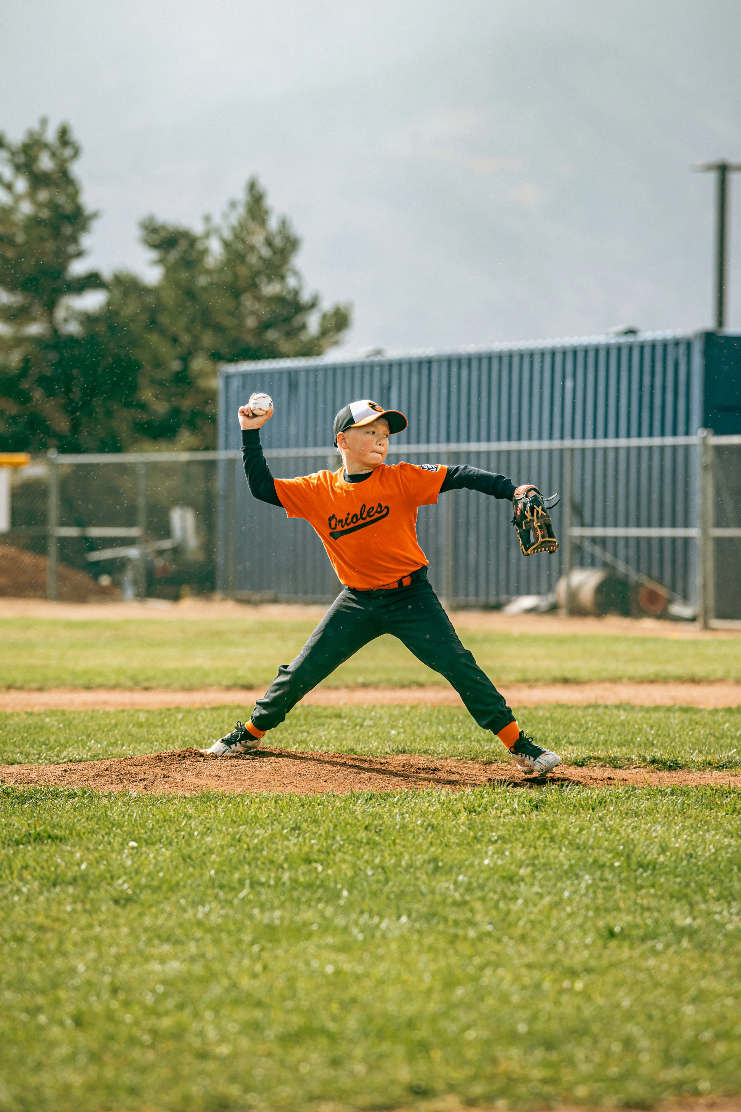A young boy in a baseball uniform pitches a ball on a baseball field, wearing an orange jersey, black pants, and a baseball glove.
