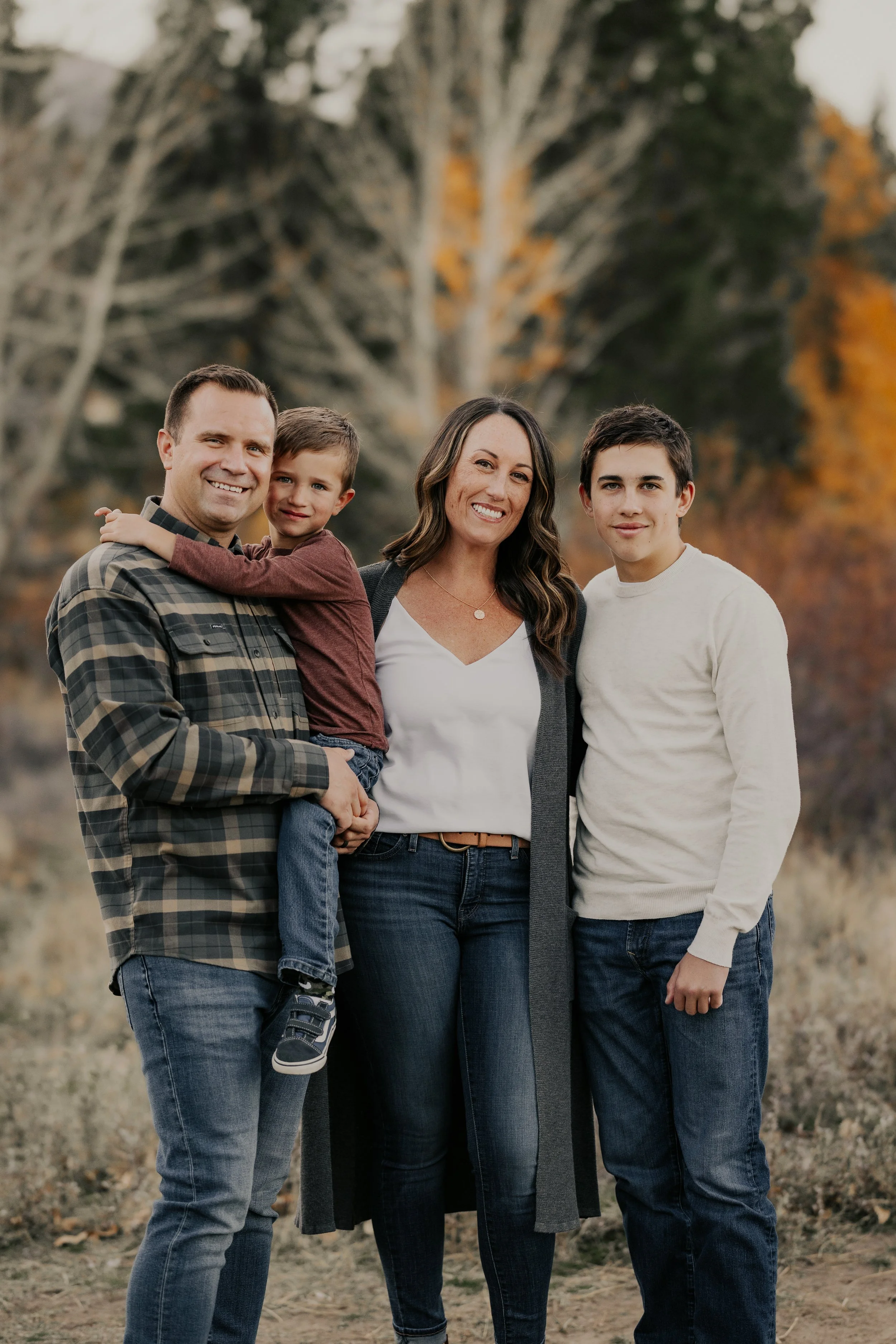 Family of four, including father, mother, and two sons, standing outdoors in fall with trees in the background, smiling at the camera.