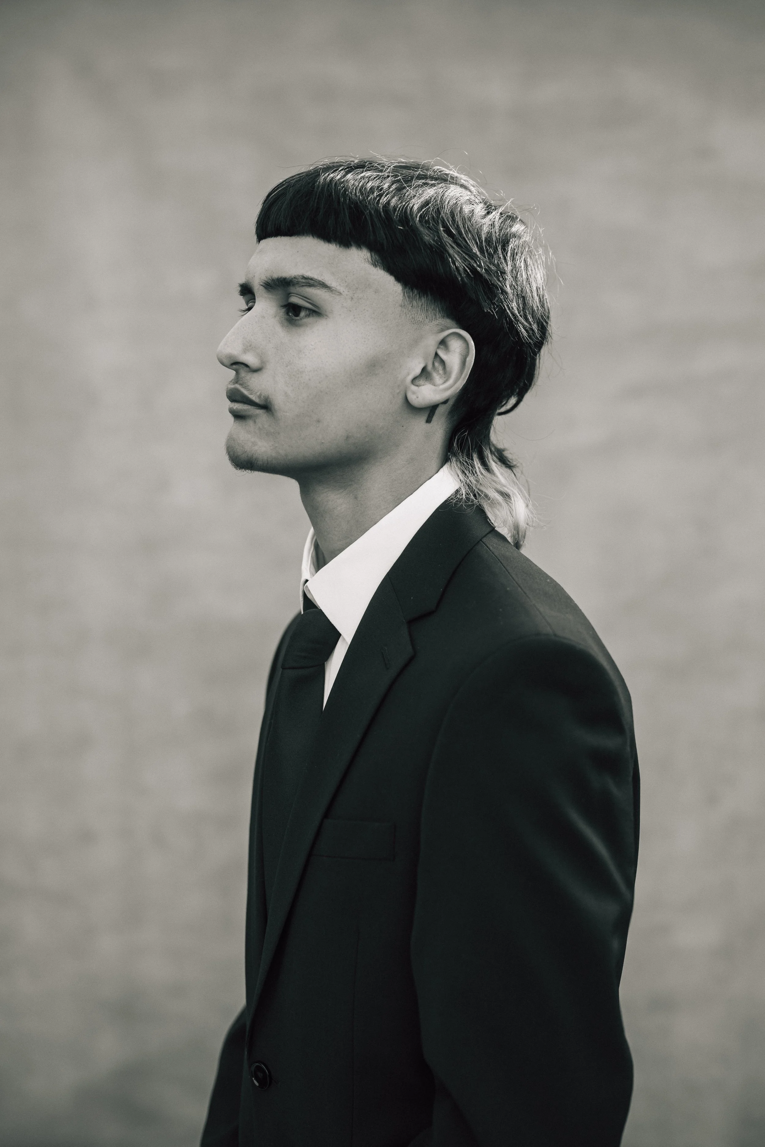A young man in a black suit with a white shirt, profile view, with a mullet hairstyle, minimal background