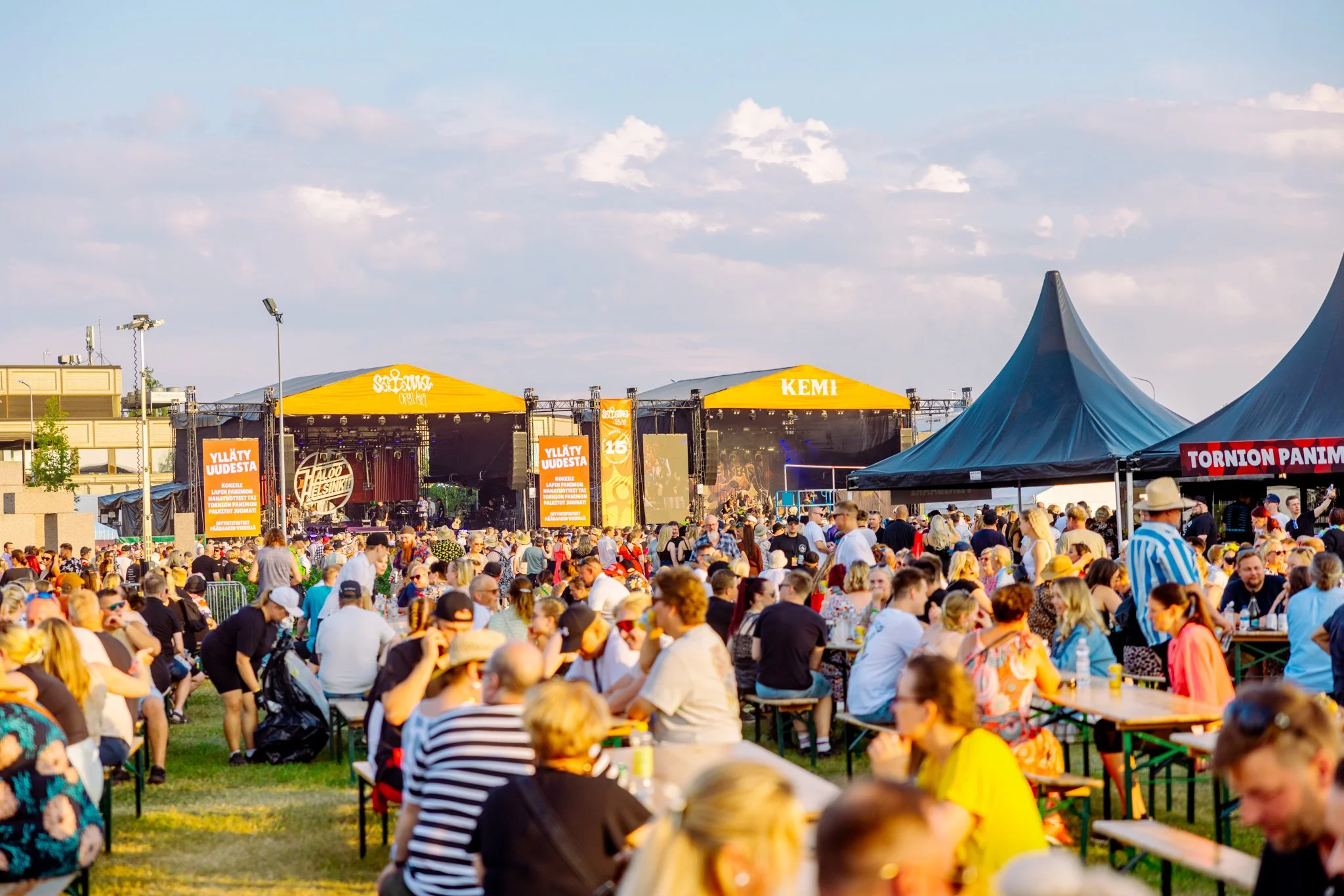 Festivaaliyleisöä istumassa pöydissä, sekä seisomassa kahden suuren festivaalilavan edessä Satama Open Airissa, Kemissä.