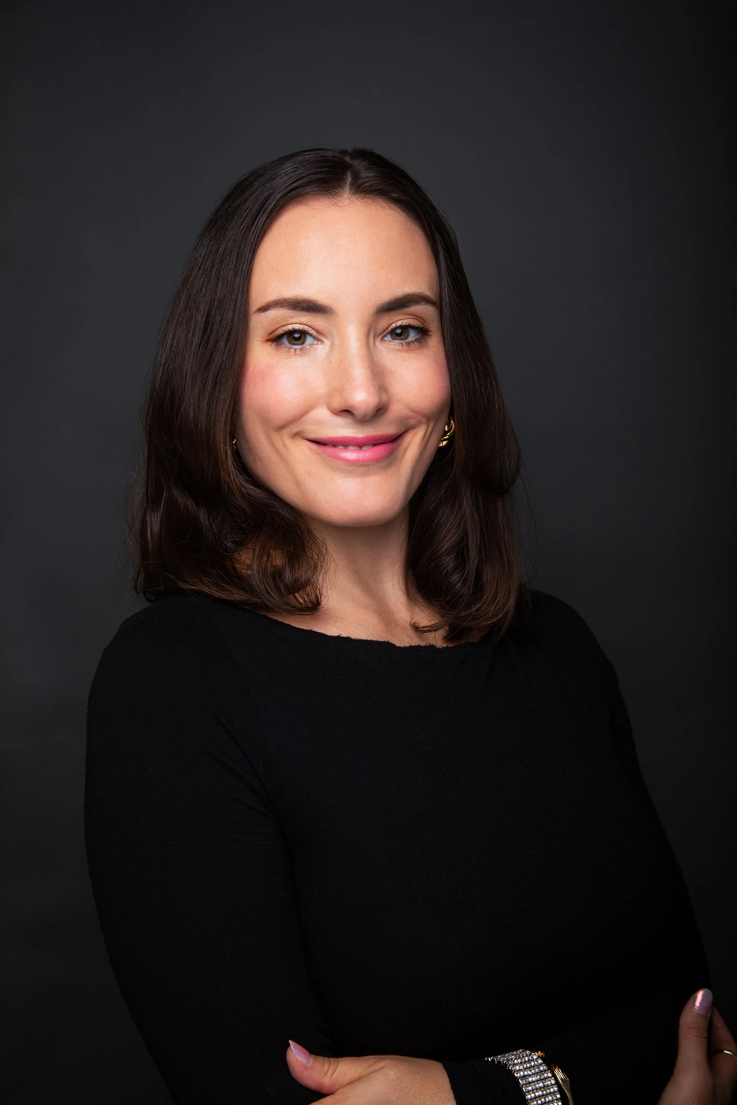 A woman with shoulder-length brown hair, wearing a black top, smiling with arms crossed against a dark background.