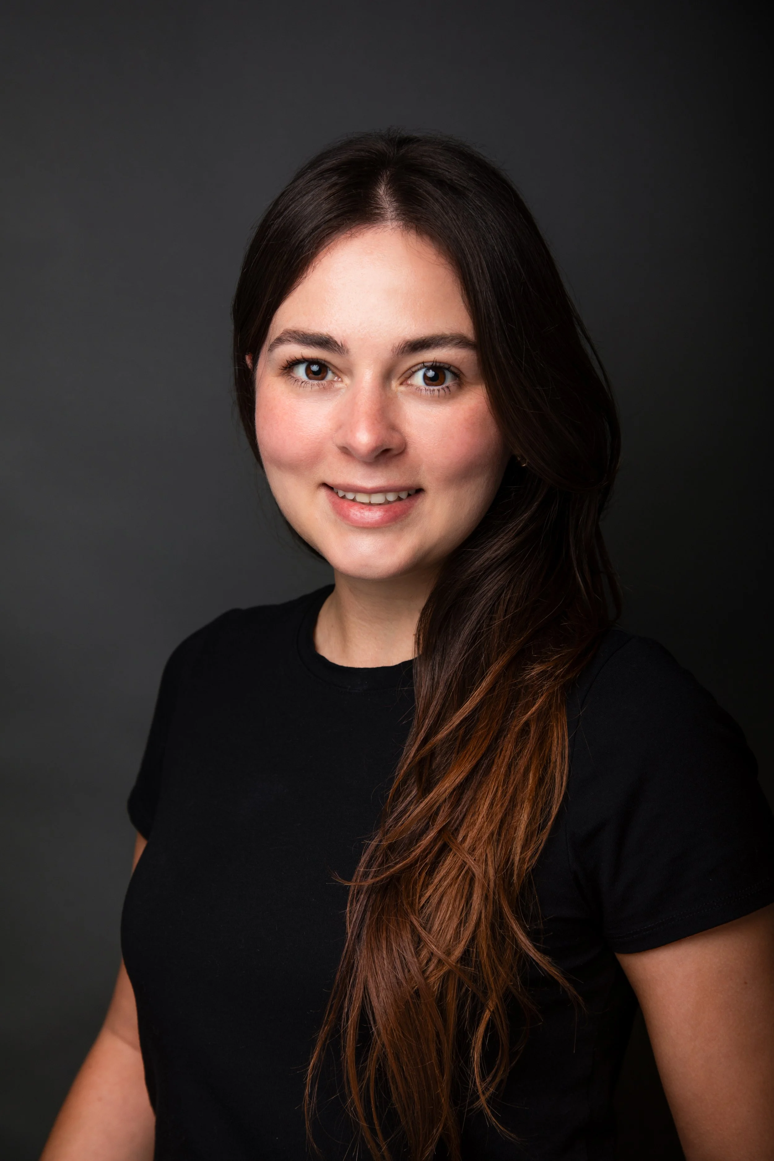 A young woman with long, dark brown hair and a friendly smile, wearing a black shirt, posed against a dark gray background.