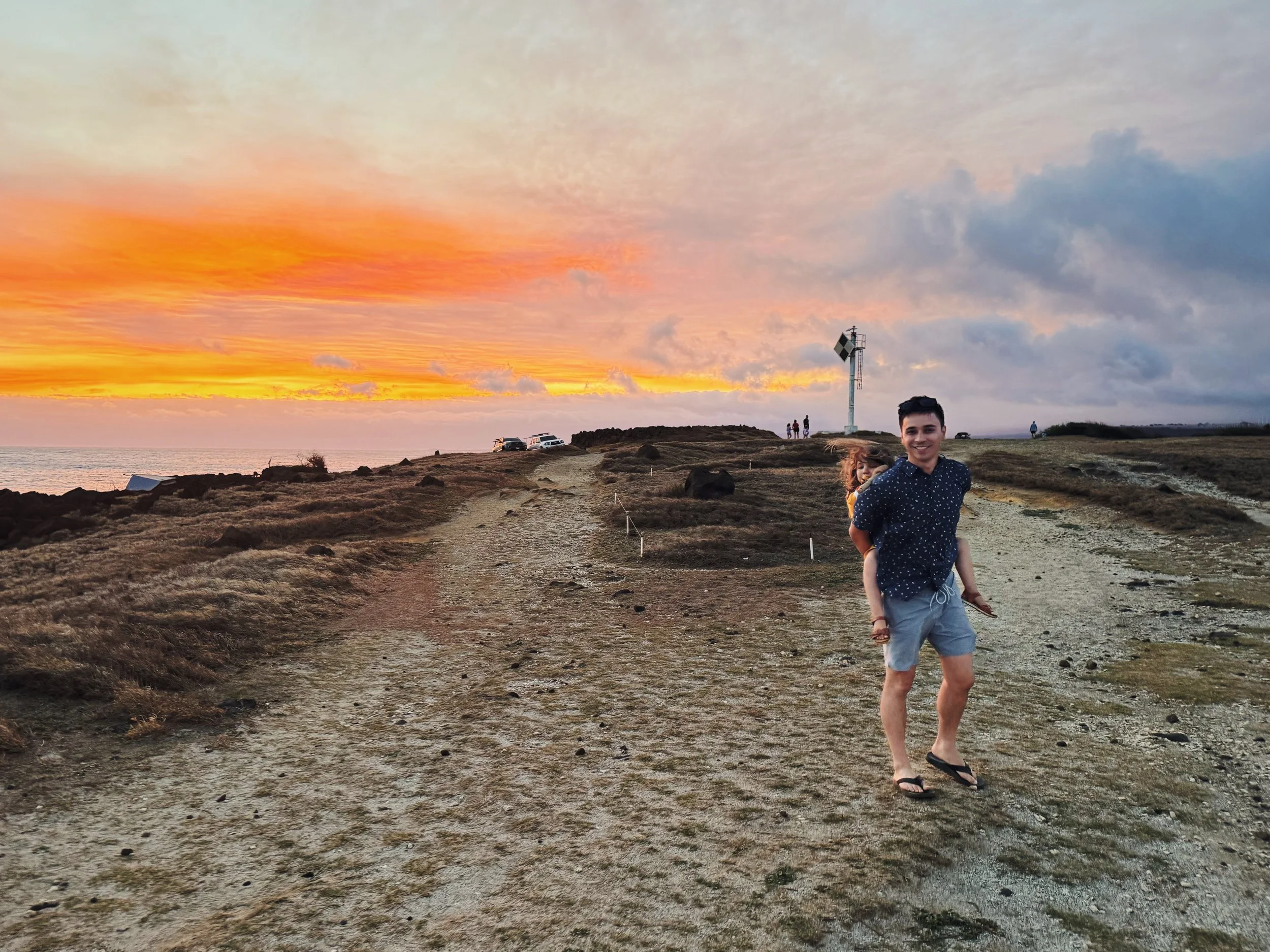 A young man carrying a girl on his back, walking along a dirt path at sunset near the ocean, with a lighthouse and a few other people in the background.