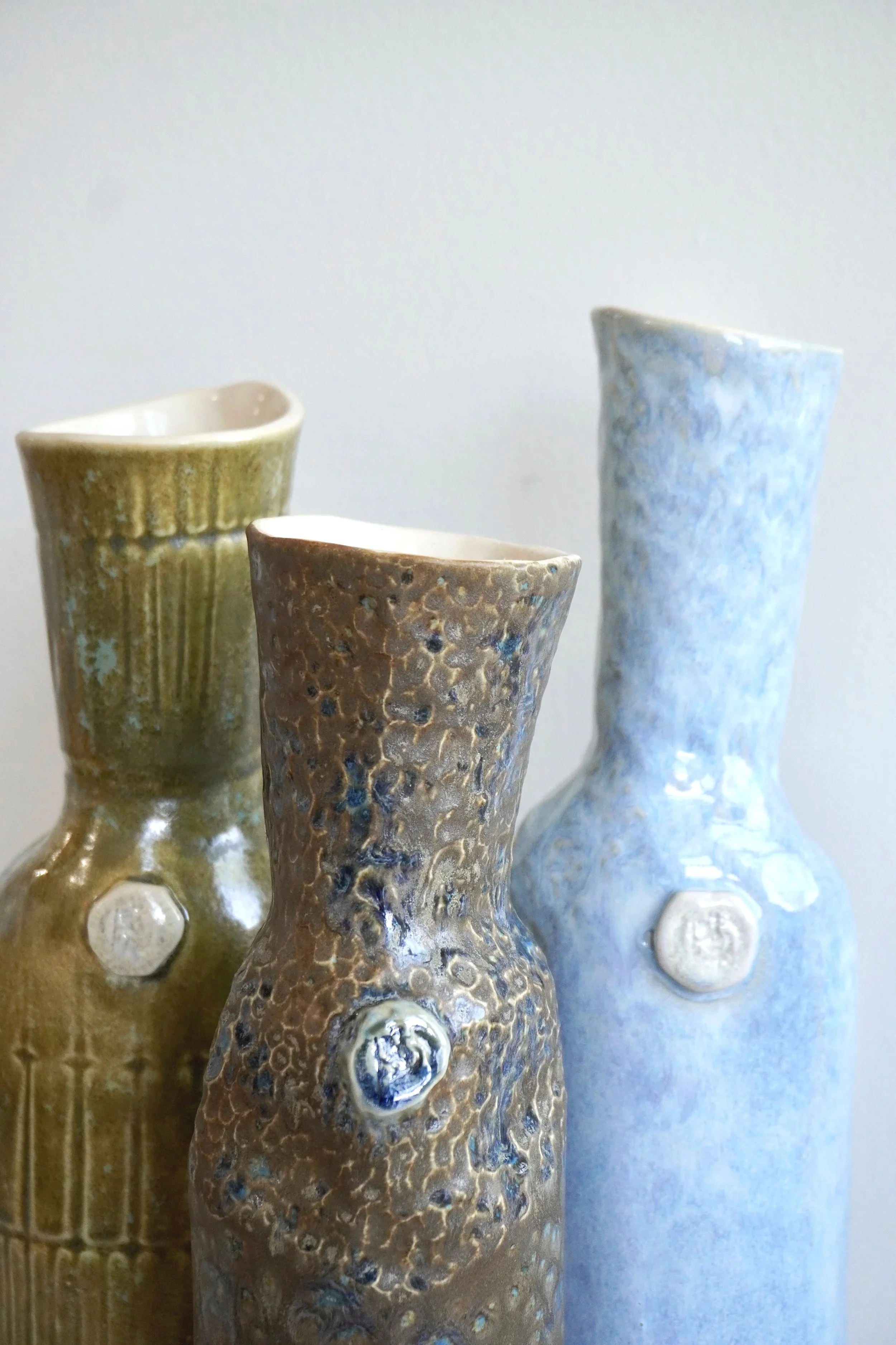 Three ceramic vases with textured surfaces in earthy tones of green, brown, and blue, displayed against a simple background.