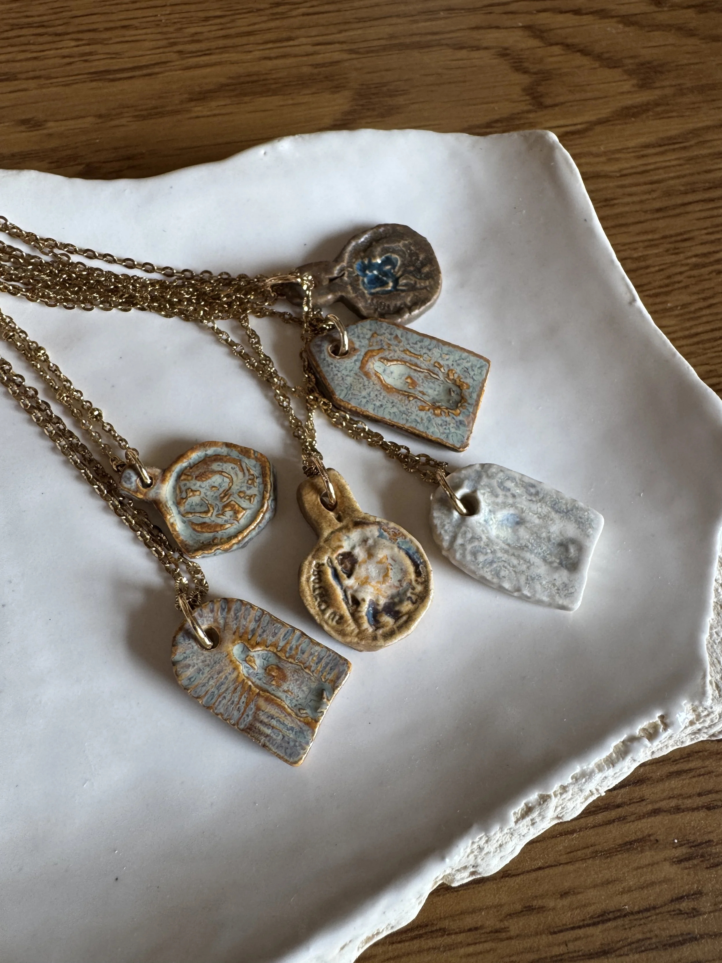 Arranged jewelry with multiple gold chains, pendants, and charms on a white plate with a rough edge, placed on a wooden surface.