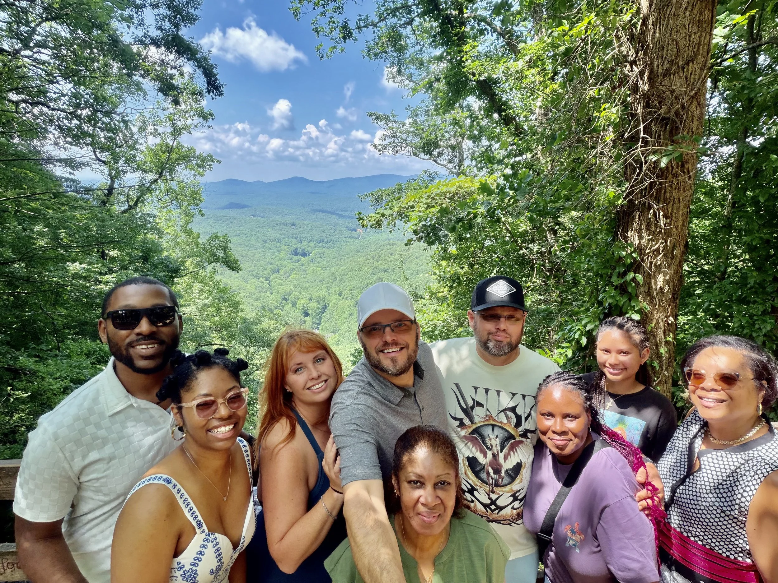 A group of diverse people smiling and posing for a photo outdoors in a wooded area with mountains and blue sky in the background.