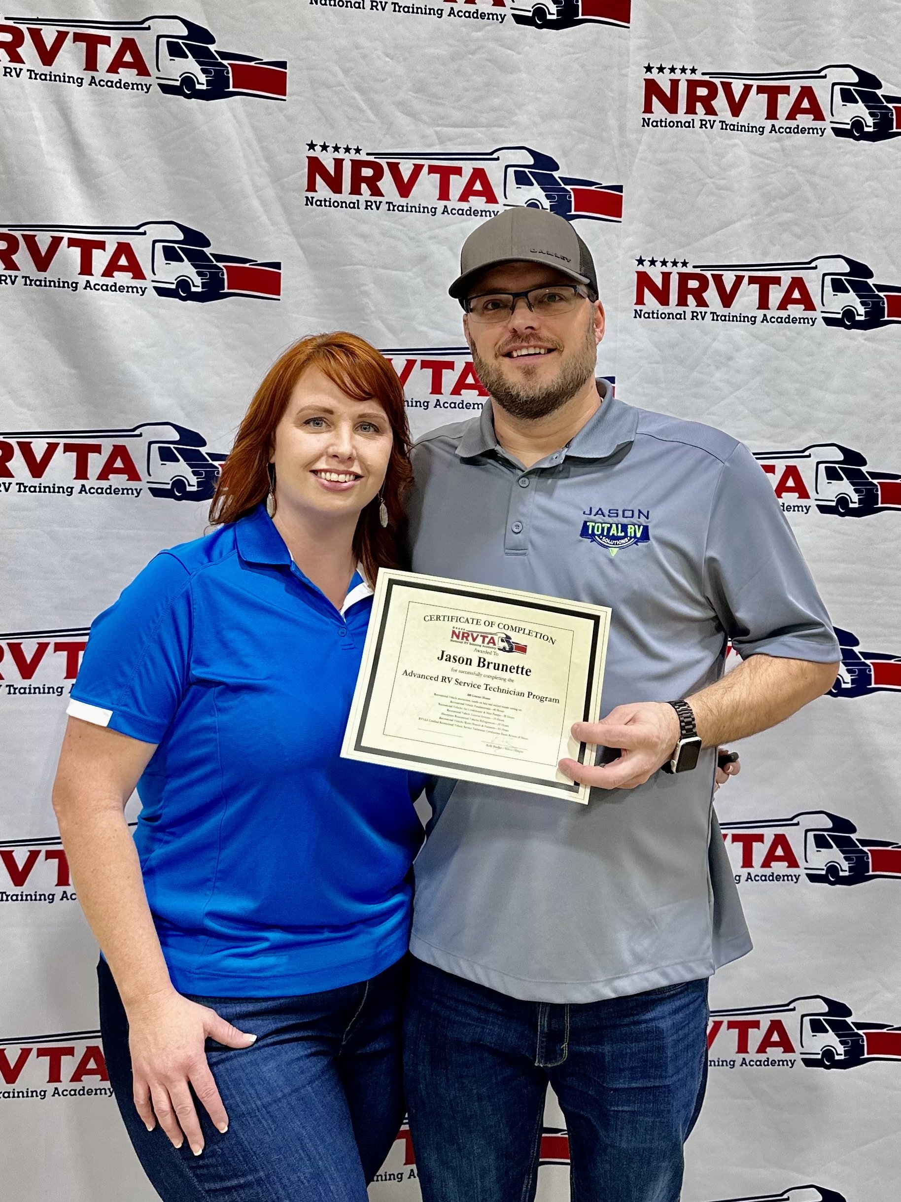 A woman and a man standing in front of a NRVTA (National RV Training Academy) banner, holding a certificate of completion. The woman has red hair and is wearing a blue shirt, while the man has a beard, glasses, a gray cap, and a gray shirt with the name tag "Jason" and the logo "Total RV."