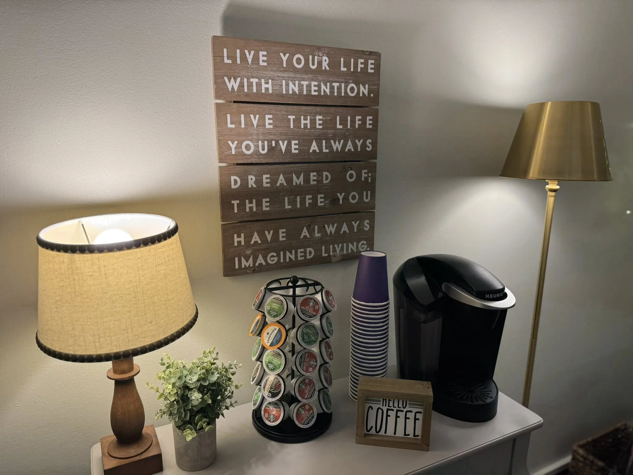 A coffee station with a Keurig machine, a sign that says "Hello Coffee," a coffee pod holder, a small plant, a bookshelf sign with a motivational quote, and two lamps on a white table against a white wall.