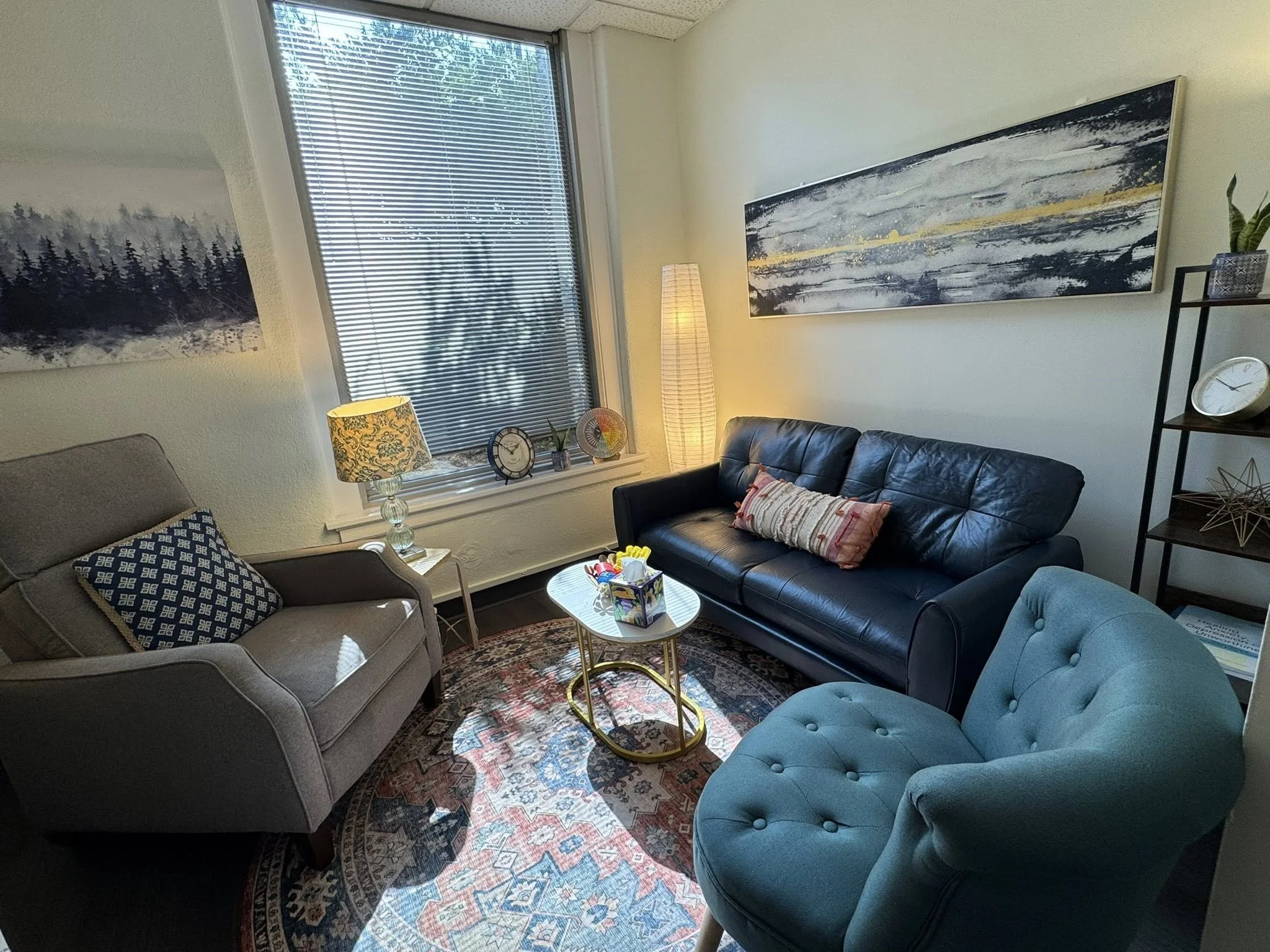 A cozy living room featuring a gray armchair with a patterned pillow, a black leather sofa with a decorative pillow, a light blue tufted armchair, and a round coffee table with art supplies on it. There is a window with blinds, a lamp, and artwork on the walls, including a large horizontal piece above the sofa and a smaller piece to the left.