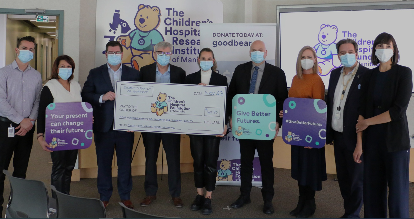 Group of nine people standing together indoors, holding signs and a large check, at the Children's Hospital Foundation of Manitoba charity event, all wearing face masks.
