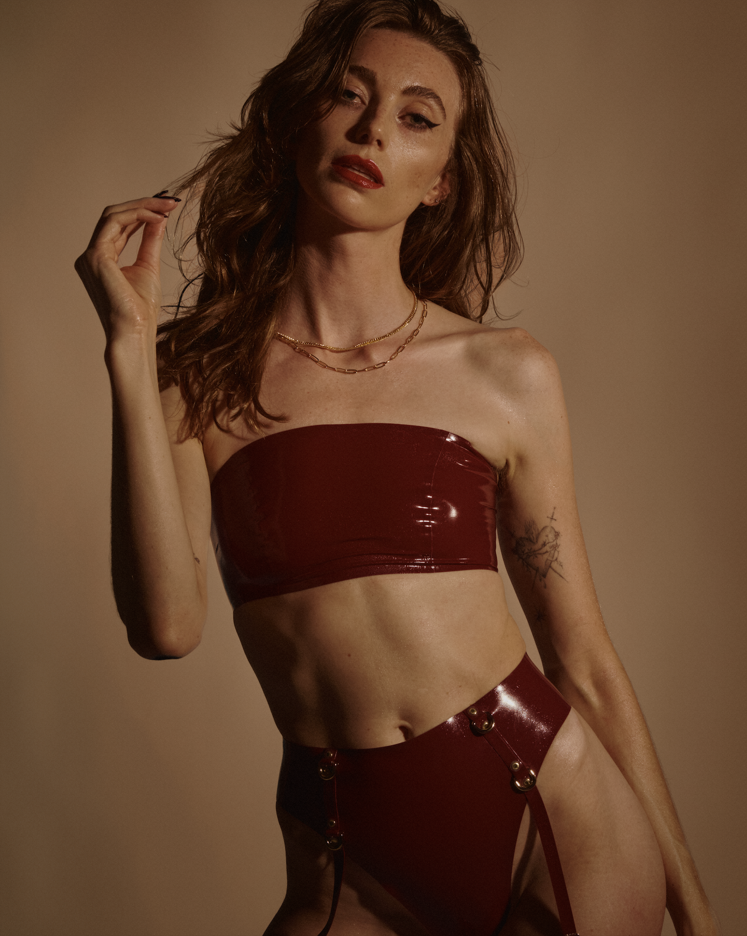 A woman with red hair and makeup wearing a strapless shiny red leather bandeau top and matching high-waisted bottoms with suspenders, against a plain brown background.