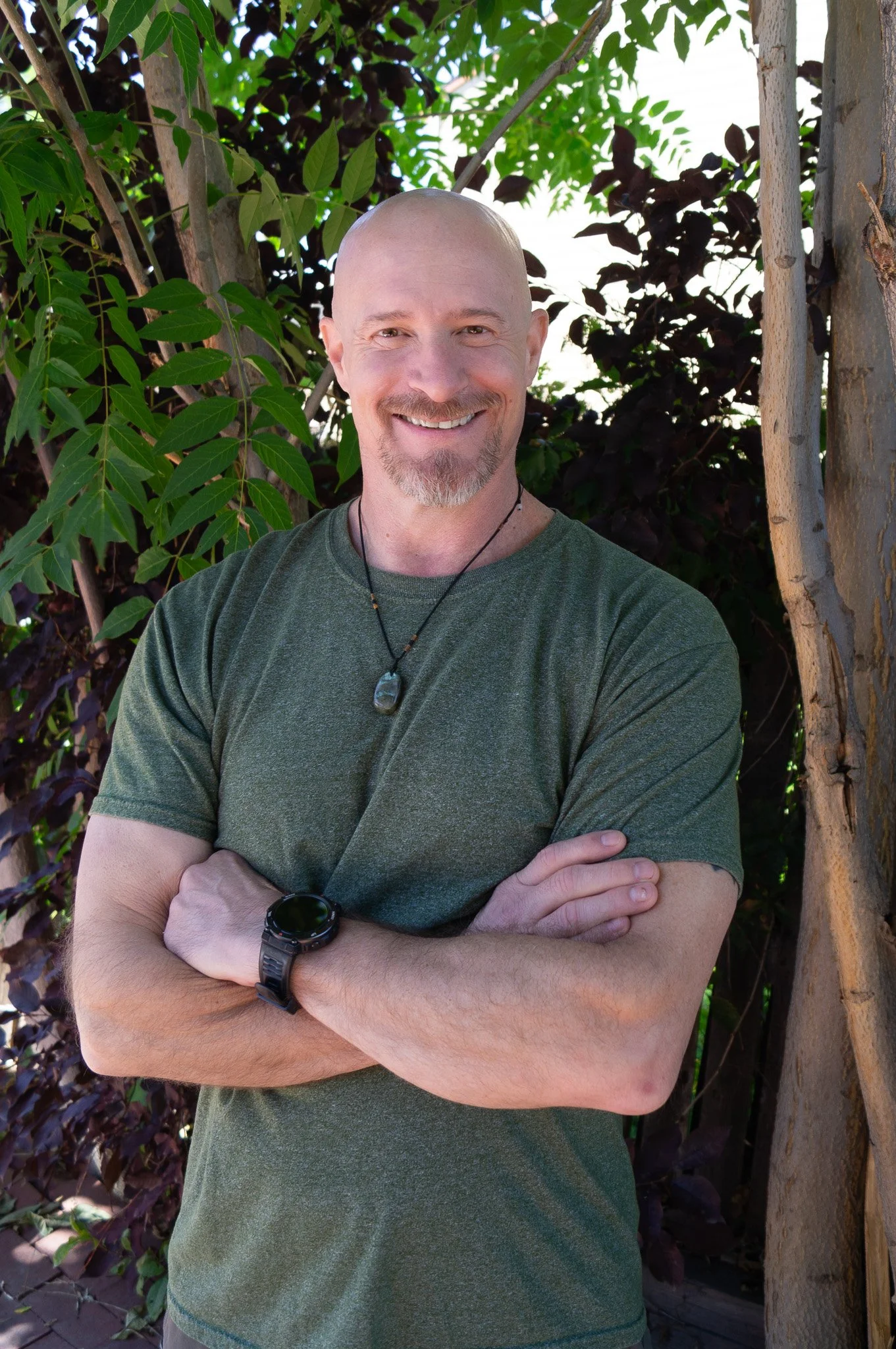 A smiling man with a bald head and beard wearing a green t-shirt, necklace, watch, and standing outdoors among trees and foliage.