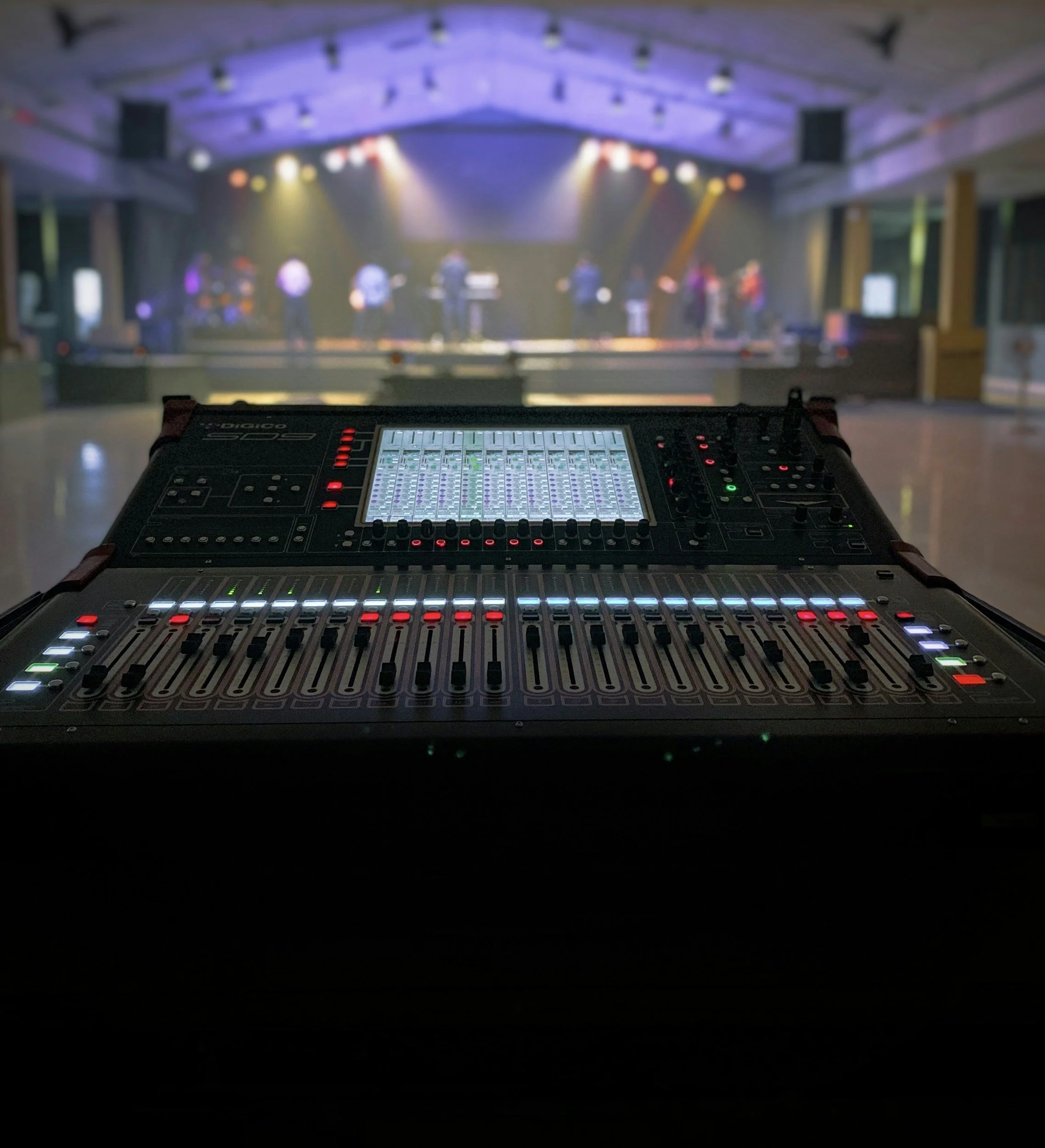 A professional audio mixer in the foreground with a blurred stage and band performance in the background.