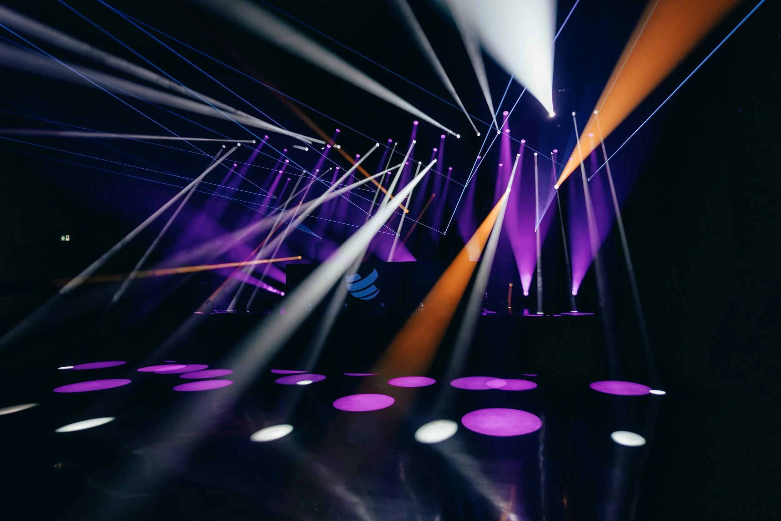 A dark stage with multiple colorful spotlights and laser lights projecting beams and circles in purple, white, and orange.