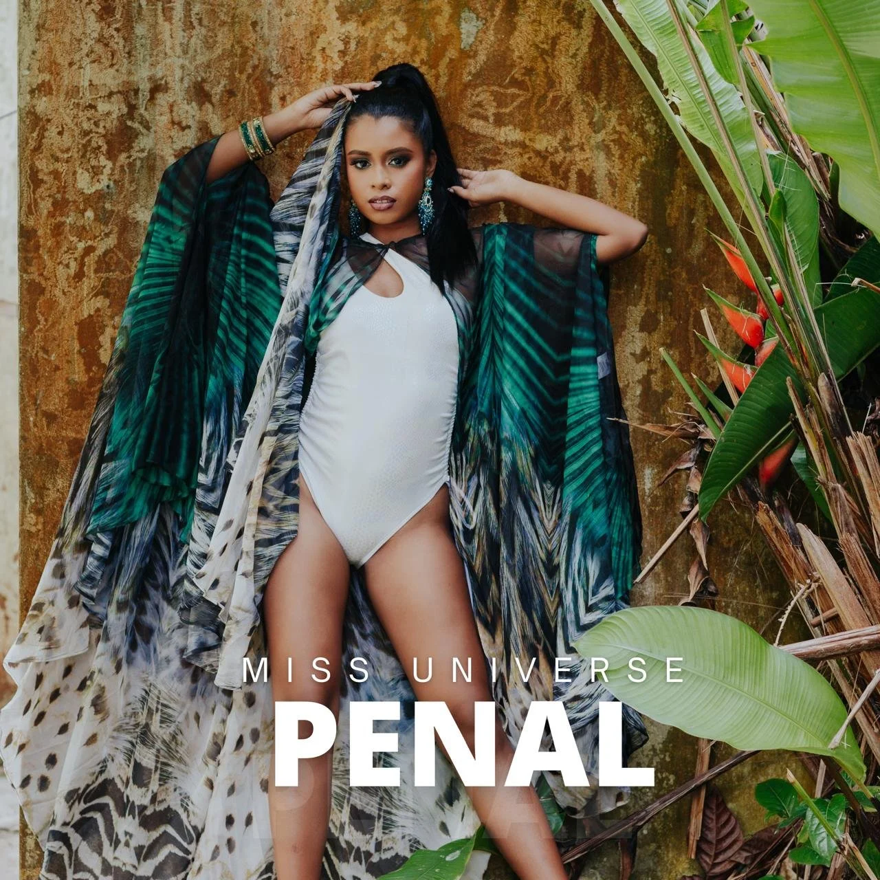 Miss Universe Penal contestant wearing a white swimsuit and a flowing patterned cover-up, standing against tropical plants and a textured brown background.