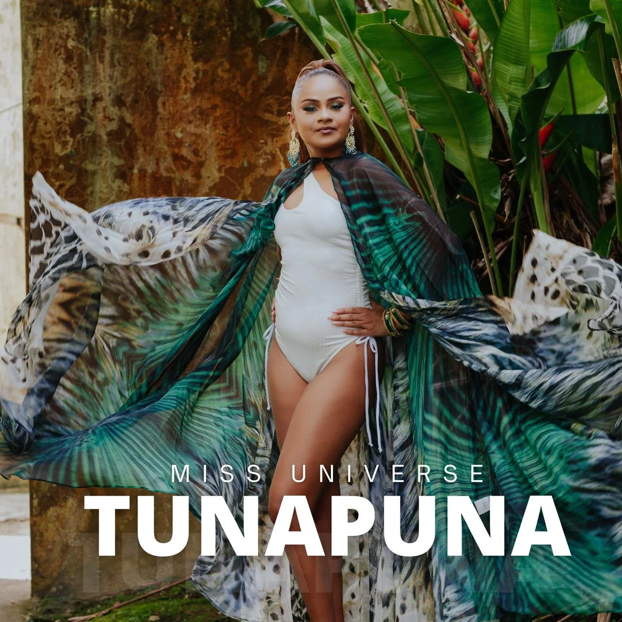 Miss Universe Tunapuna contestant in a white swimsuit with a multicolored, flowing cover-up, standing outdoors with tropical plants in the background.