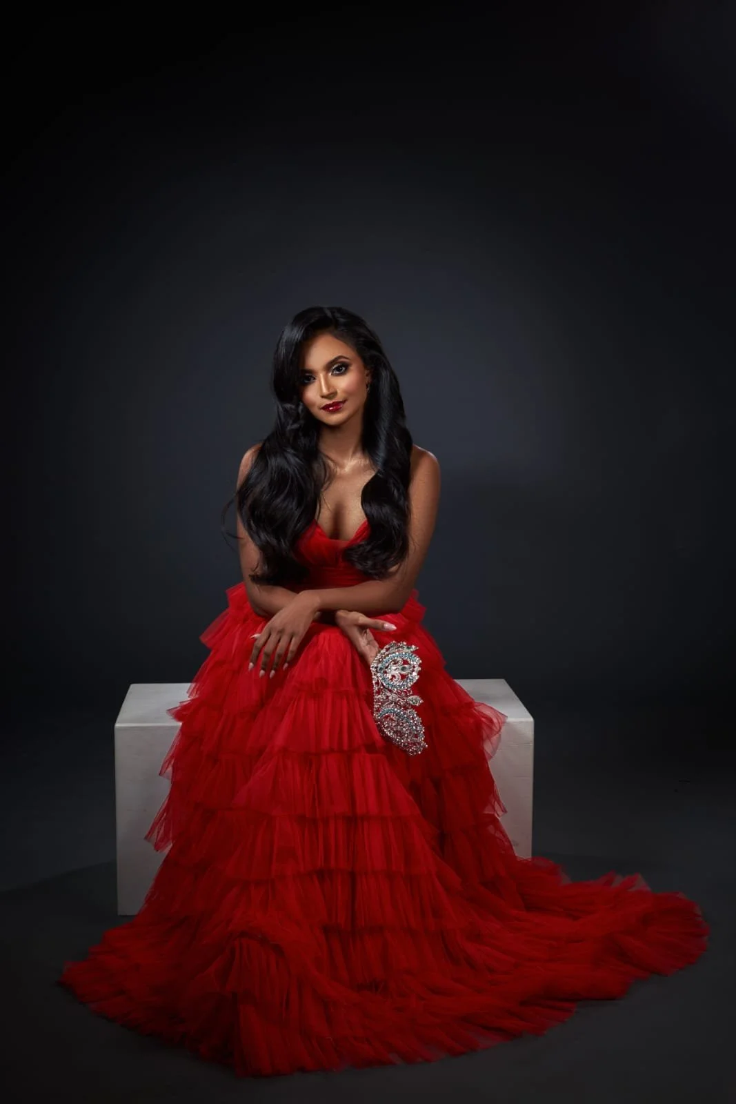 A woman sitting on a white block in a red gown with ruffles, holding a silver beaded clutch, against a dark background.