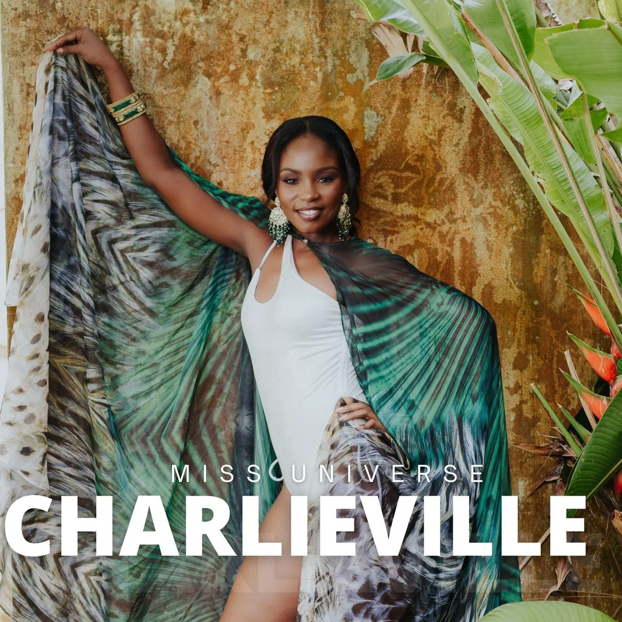 A woman poses confidently with one arm raised, wearing a white dress and a flowing, multicolored, patterned shawl. She has dark hair, large earrings, and bracelets, standing against a textured, earthy wall with tropical plants nearby.