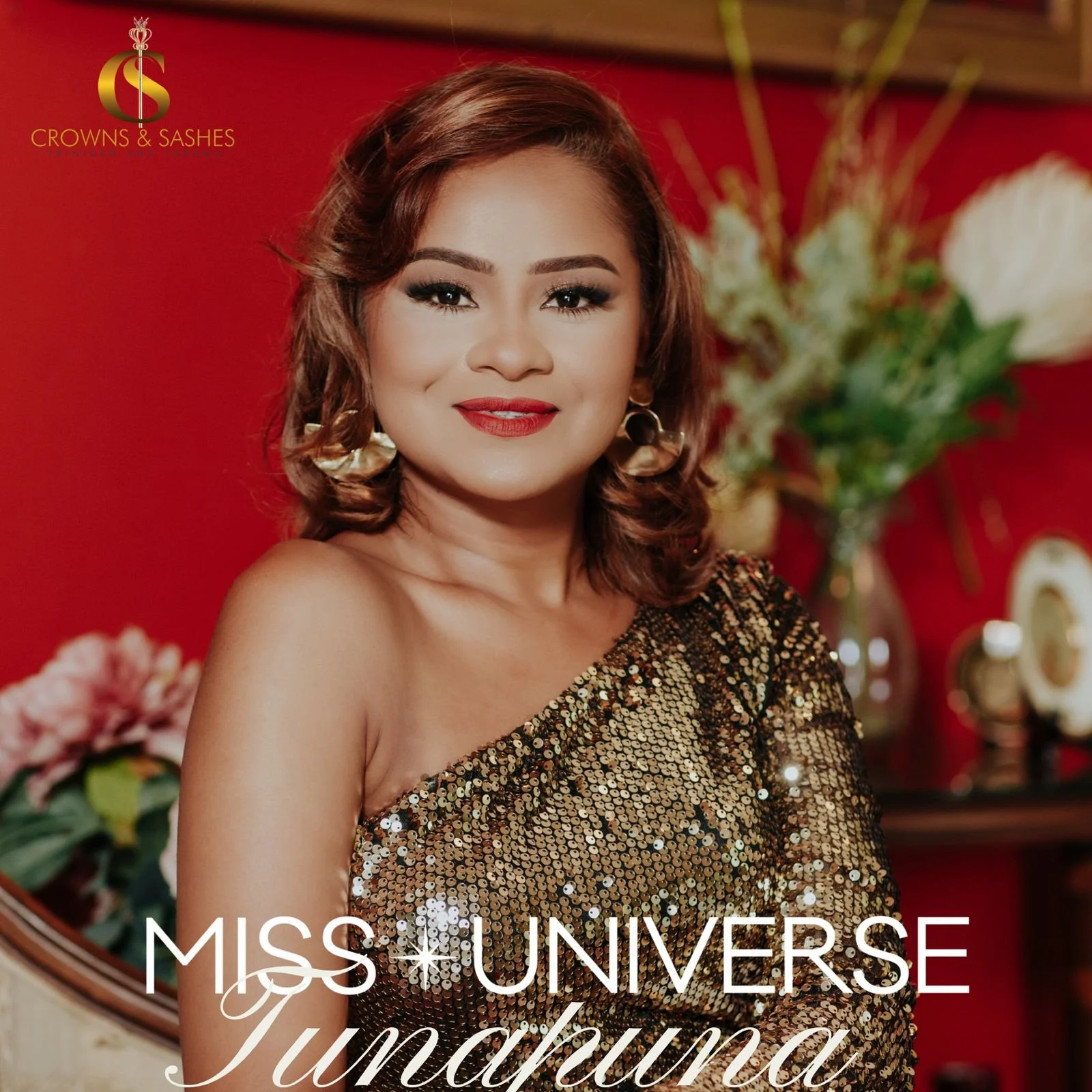 Portrait of Miss Universe Tunasura, a woman with shoulder-length wavy auburn hair, wearing a gold sequined one-shoulder dress, gold earrings, and bright red lipstick, standing in a room with a red wall, flowers, and decorative items in the background.