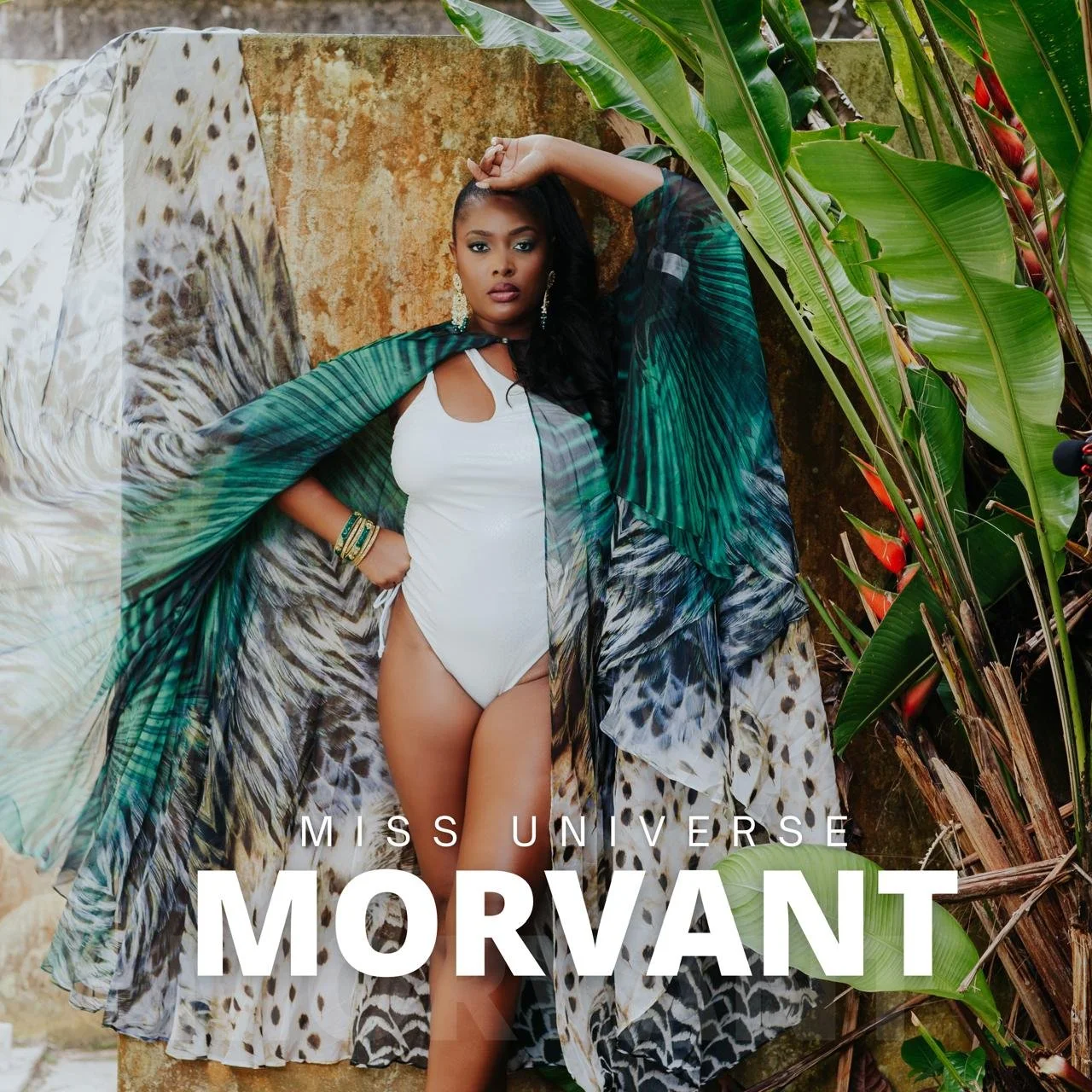 A woman dressed in a white swimsuit and a colorful outer cape, posing against a rustic wall with lush green tropical plants around her. The text on the image reads 'Miss Universe Morvant'.