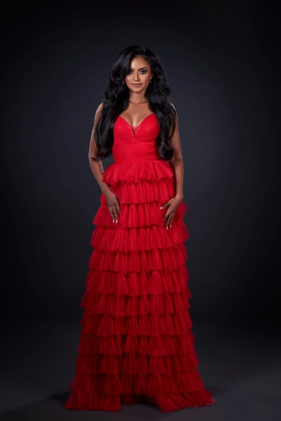 A woman with long black hair wearing a red, multi-layered ruffle gown standing against a dark background.