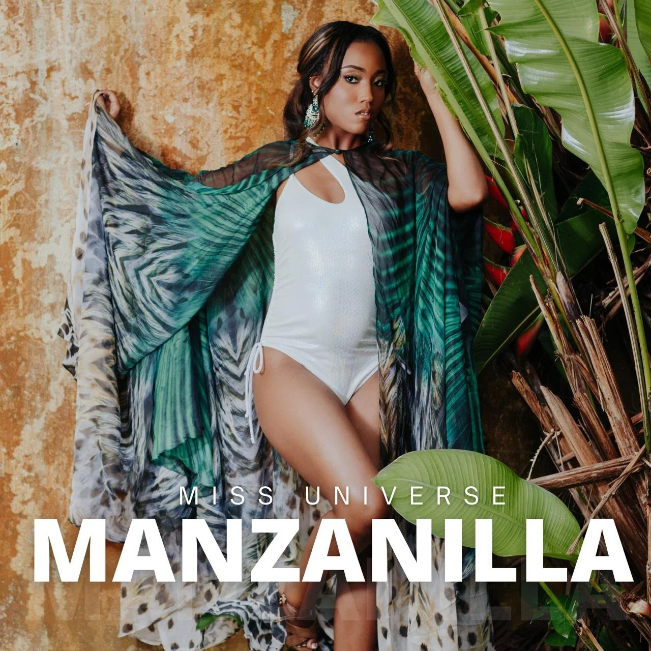 A woman posing next to lush green tropical plants and an orange textured wall, wearing a white swimsuit and a sheer, colorful sarong. The text overlay reads "Miss Universe Manzanilla."
