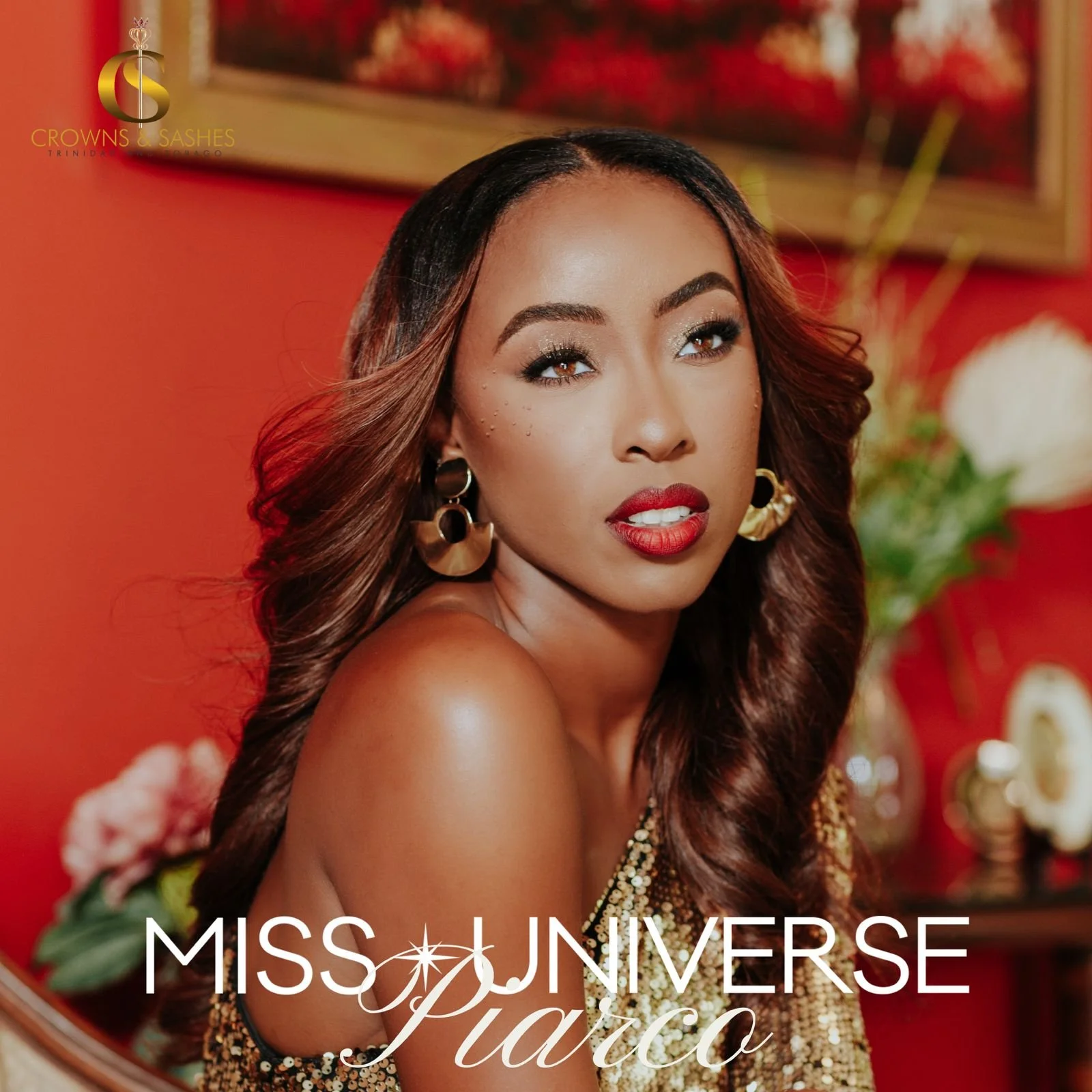 Portrait of Miss Universe Piacco with long brown hair, red lipstick, gold earrings, and glittery gold dress, in a room with red walls, floral arrangements, and framed artwork in the background.