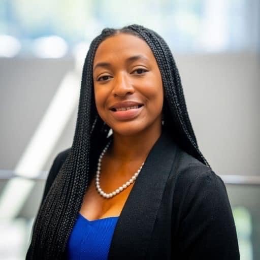 Portrait of a woman with long braided hair, wearing a black blazer and a pearl necklace, smiling at the camera.