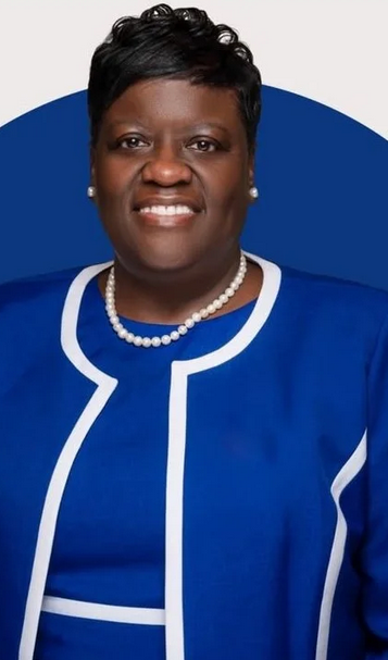 Portrait of a woman with short black hair, wearing a blue jacket with white trim, pearl necklace, and pearl earrings, smiling against a gray and blue background.