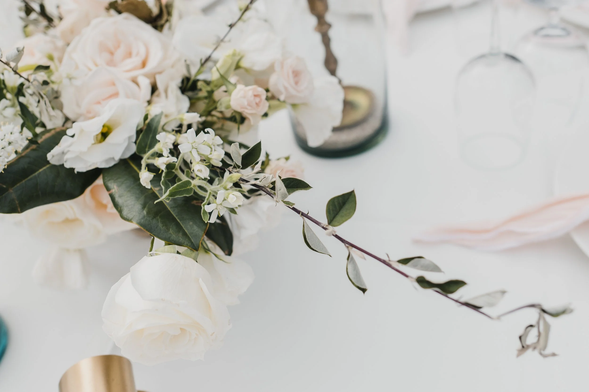 Arreglo de flores blancas y verdes sobre una mesa blanca, con copas de vino y objetos decorativos.