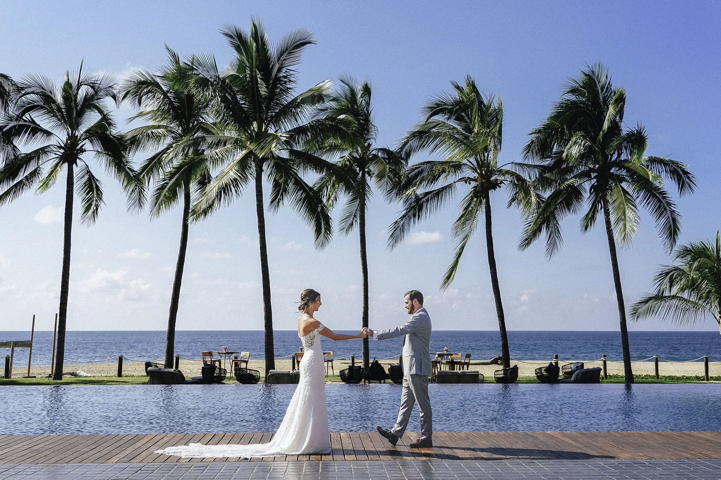 Pareja de novios bailando frente a la piscina en un lugar tropical, con palmeras y vista al mar de fondo