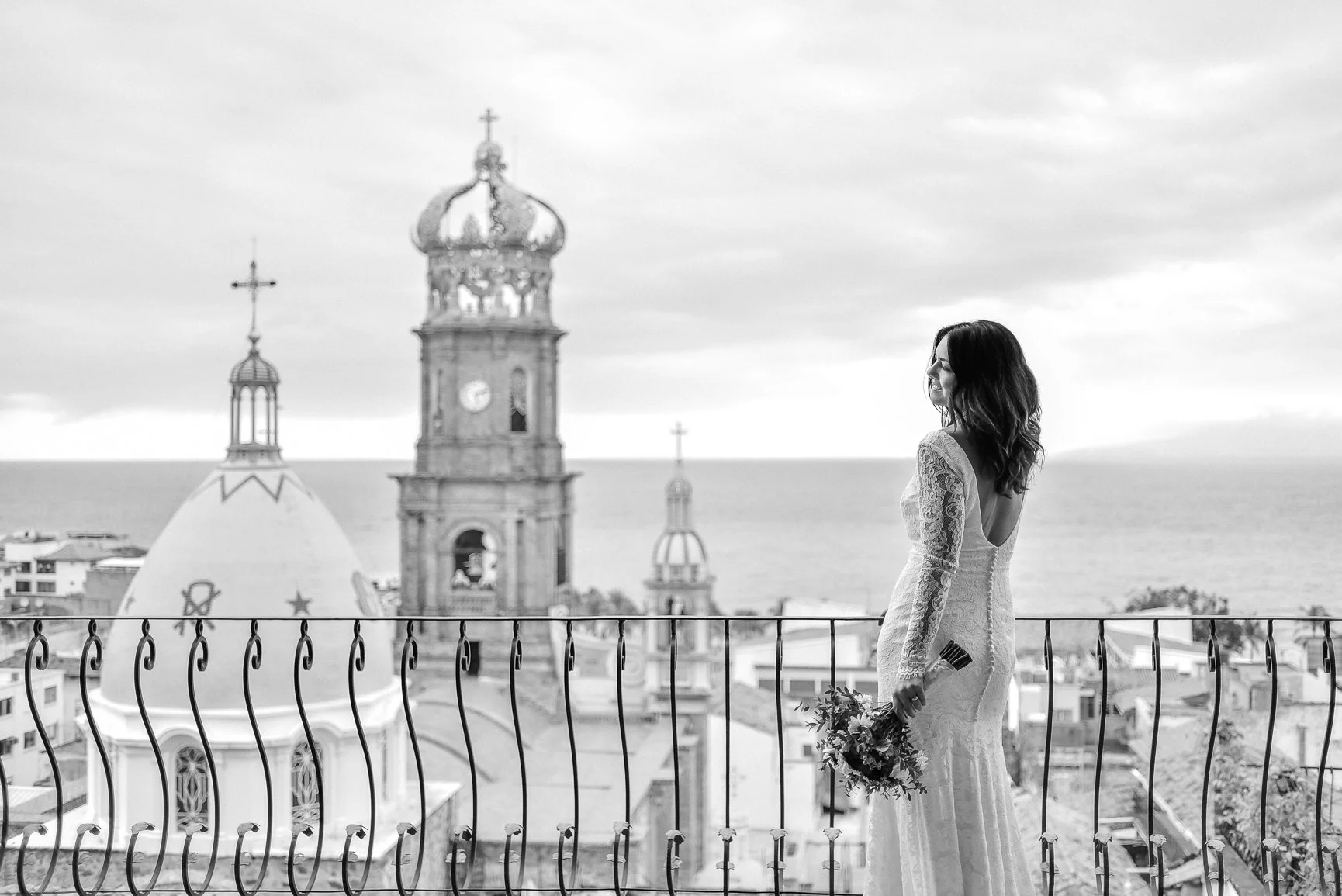 Mujer con vestido de novia sosteniendo ramo en una terraza con vista a iglesias y el mar en el fondo en un día nublado.
