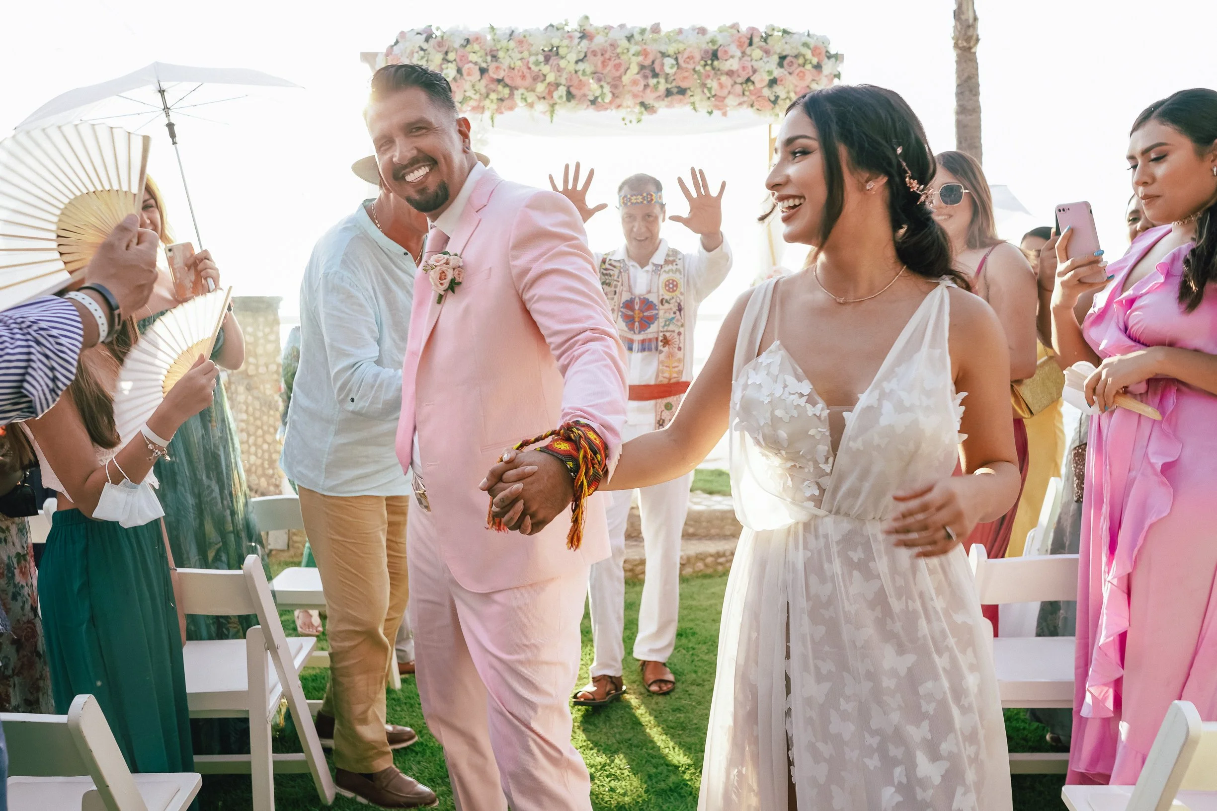 Una pareja de recién casados en una ceremonia al aire libre, rodeada de amigos celebrando y tomando fotos, con decoraciones florales y ambiente festivo.