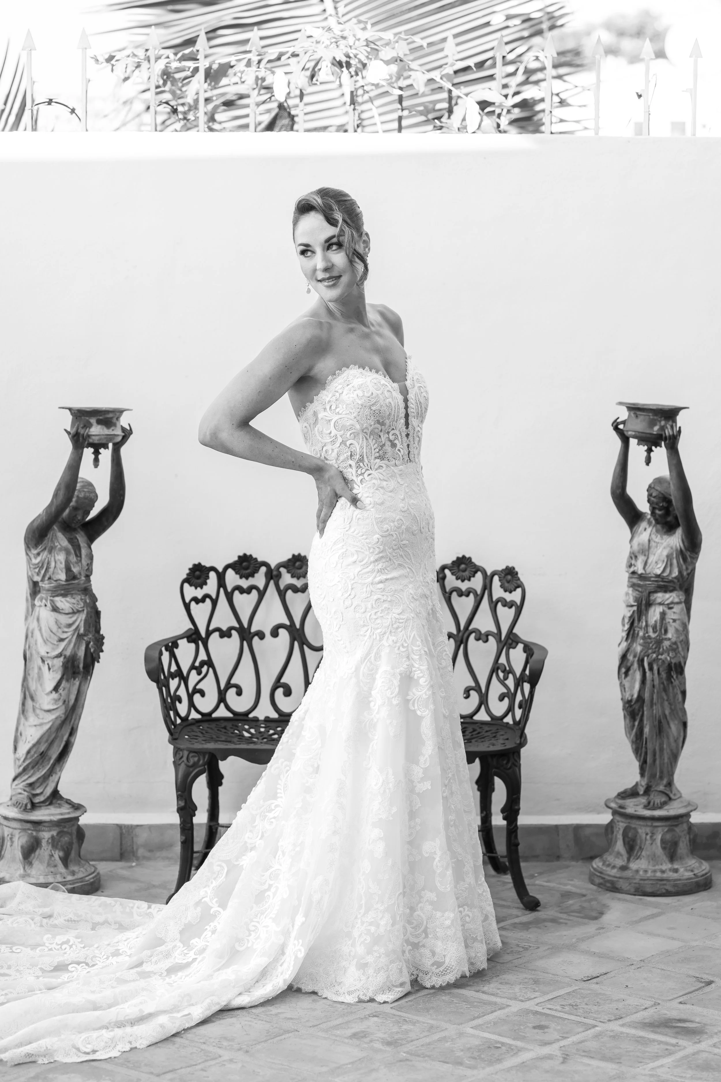 Una mujer vestida con un vestido de novia de encaje, de pie entre dos estatuas antiguas, en un entorno decorado con un banco de hierro y plantas.