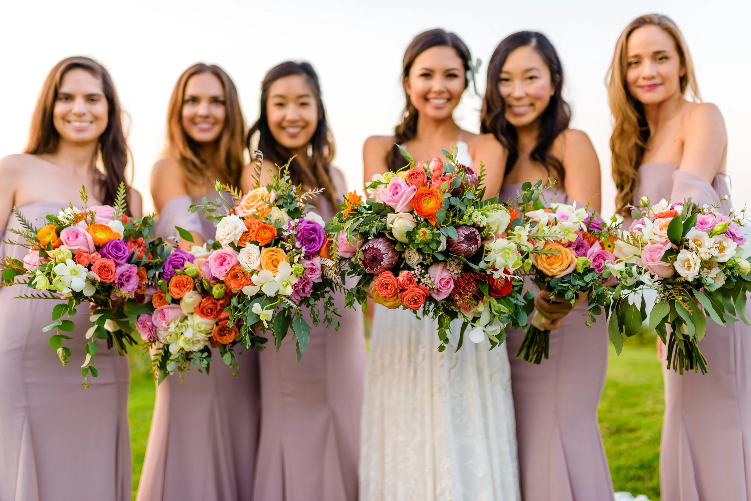 Grupo de mujeres con vestidos morados, cada una sostiene un ramo de flores coloridas, en una celebración al aire libre.