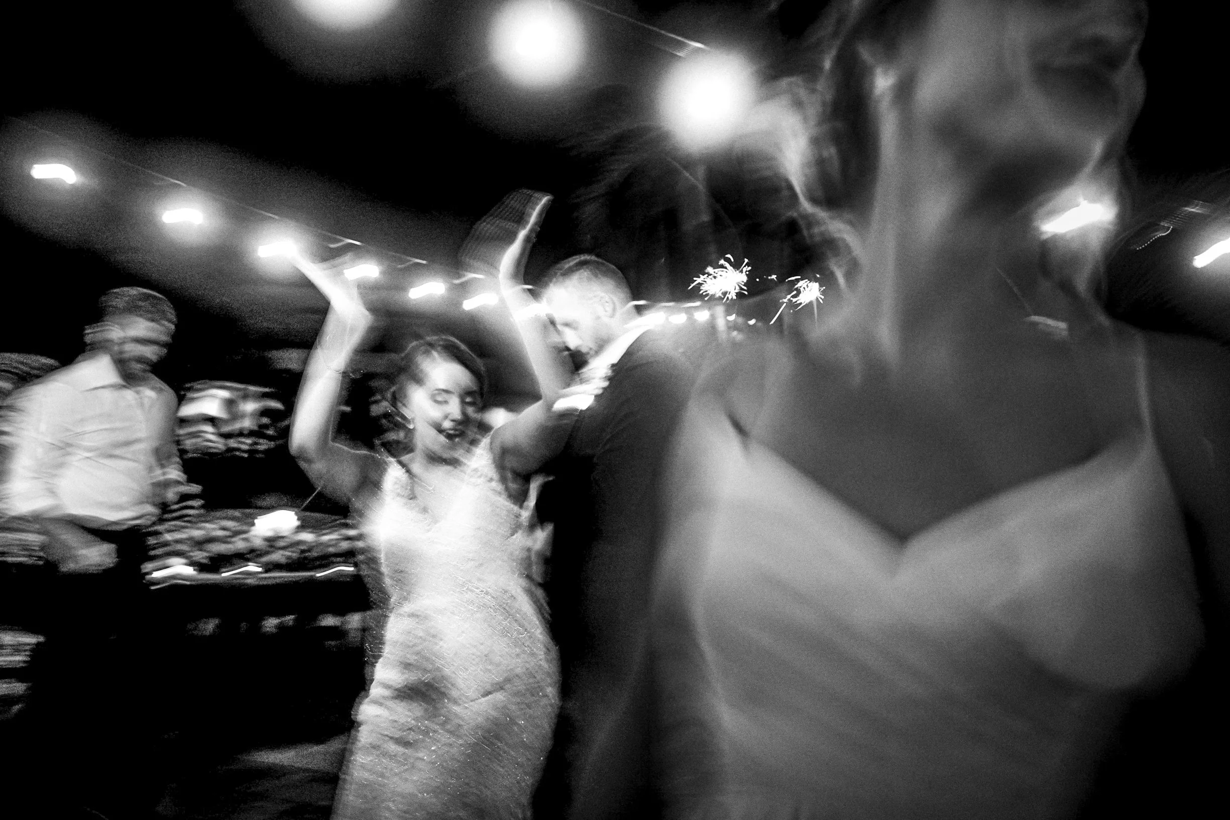 Pareja bailando en una fiesta con luces y personas en el fondo, en blanco y negro.