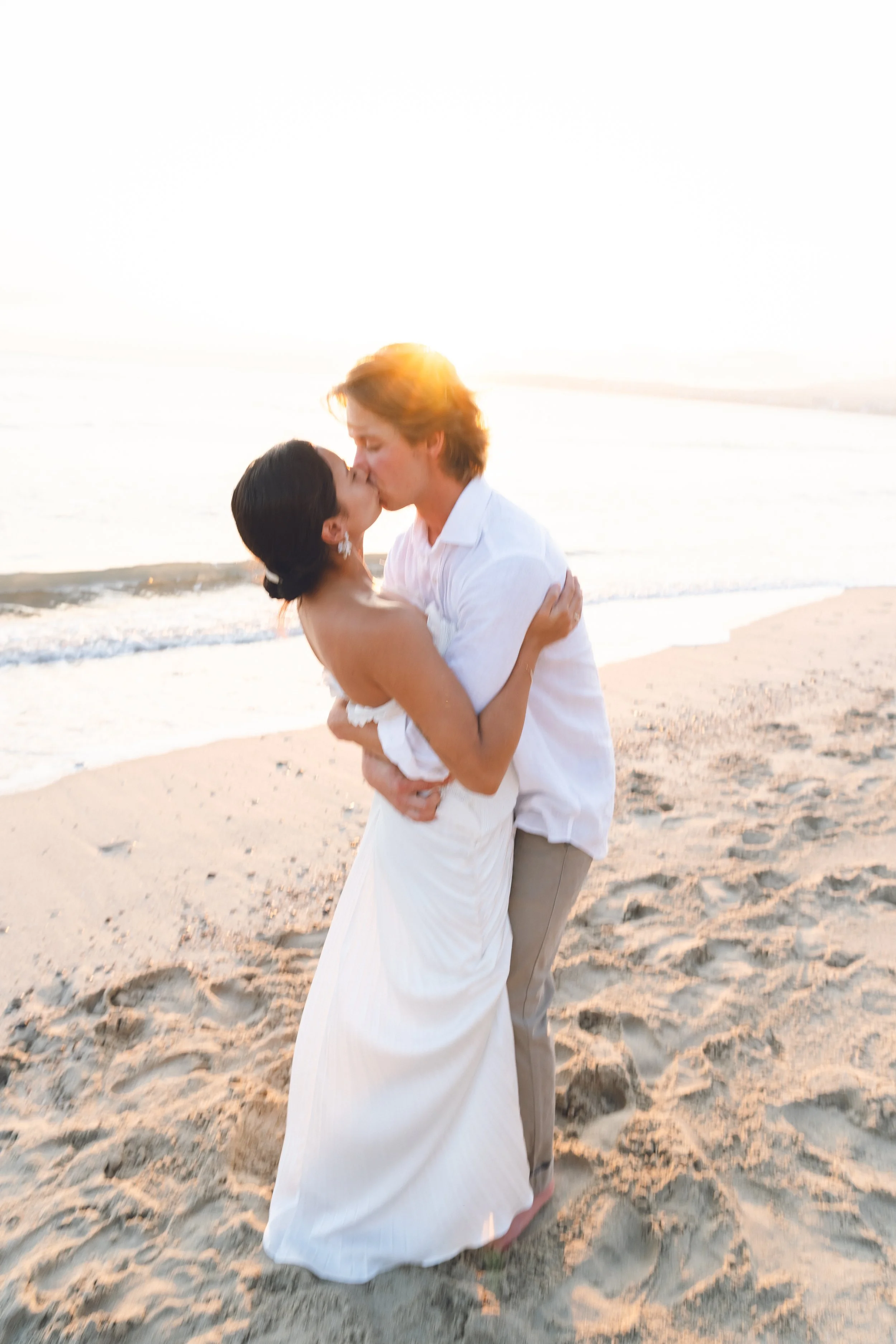 Una pareja besándose en la playa durante el atardecer, con el mar de fondo y arena a sus pies.