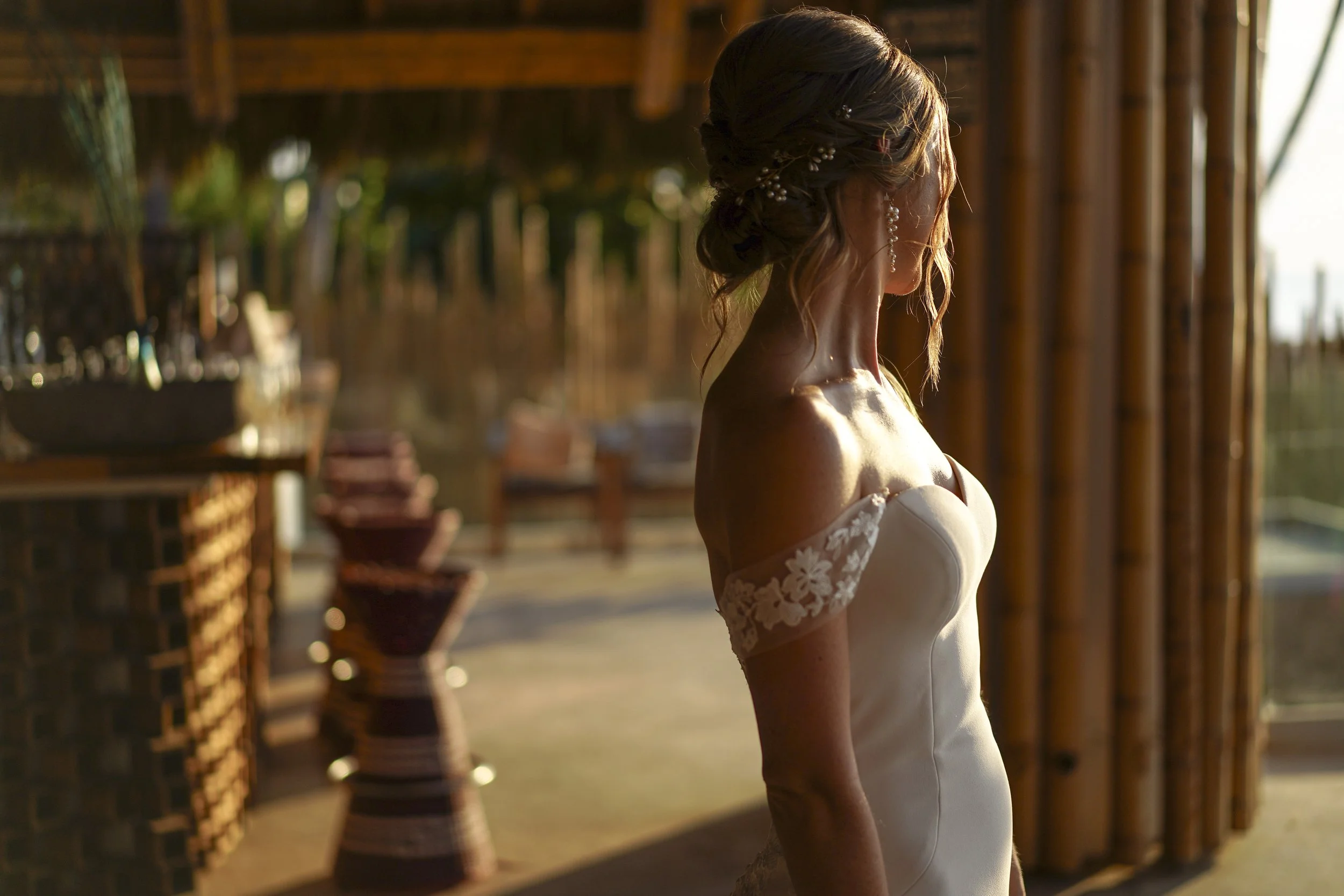 Mujer con vestido de novia de perfil en un lugar con paredes de bambú y luz cálida de atardecer.