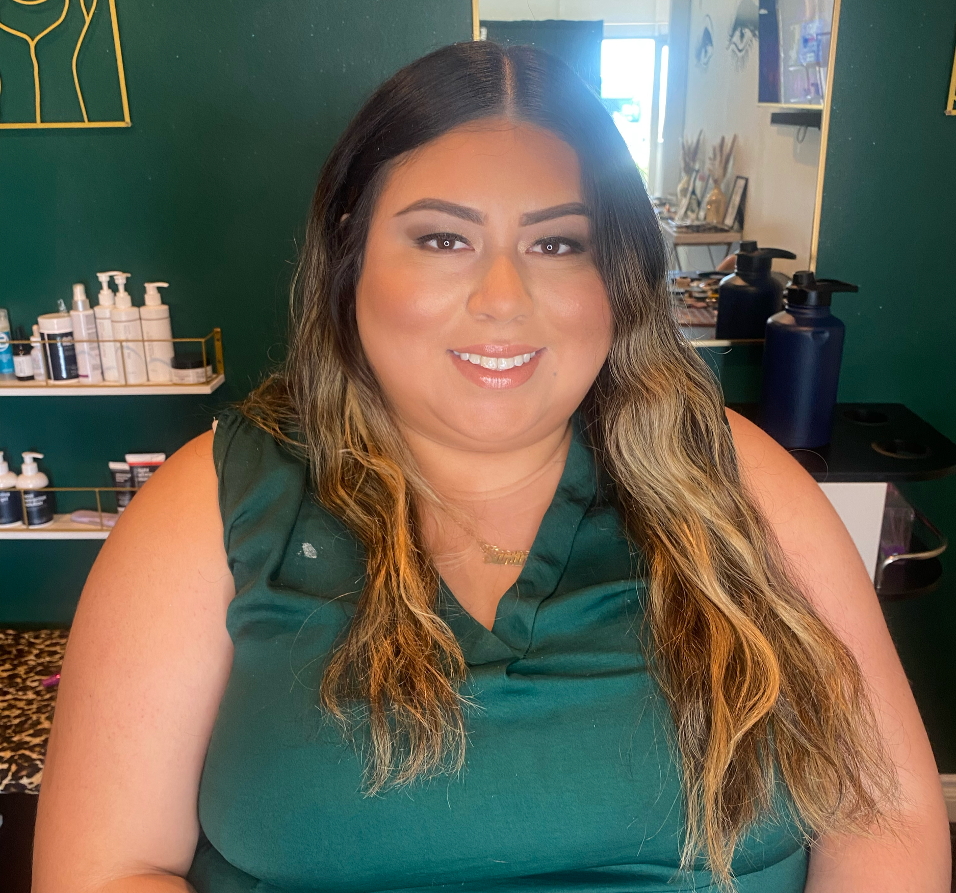 A woman with long wavy hair and makeup, smiling in a salon or store with green walls, hair care products on shelves, and a mirror in the background.