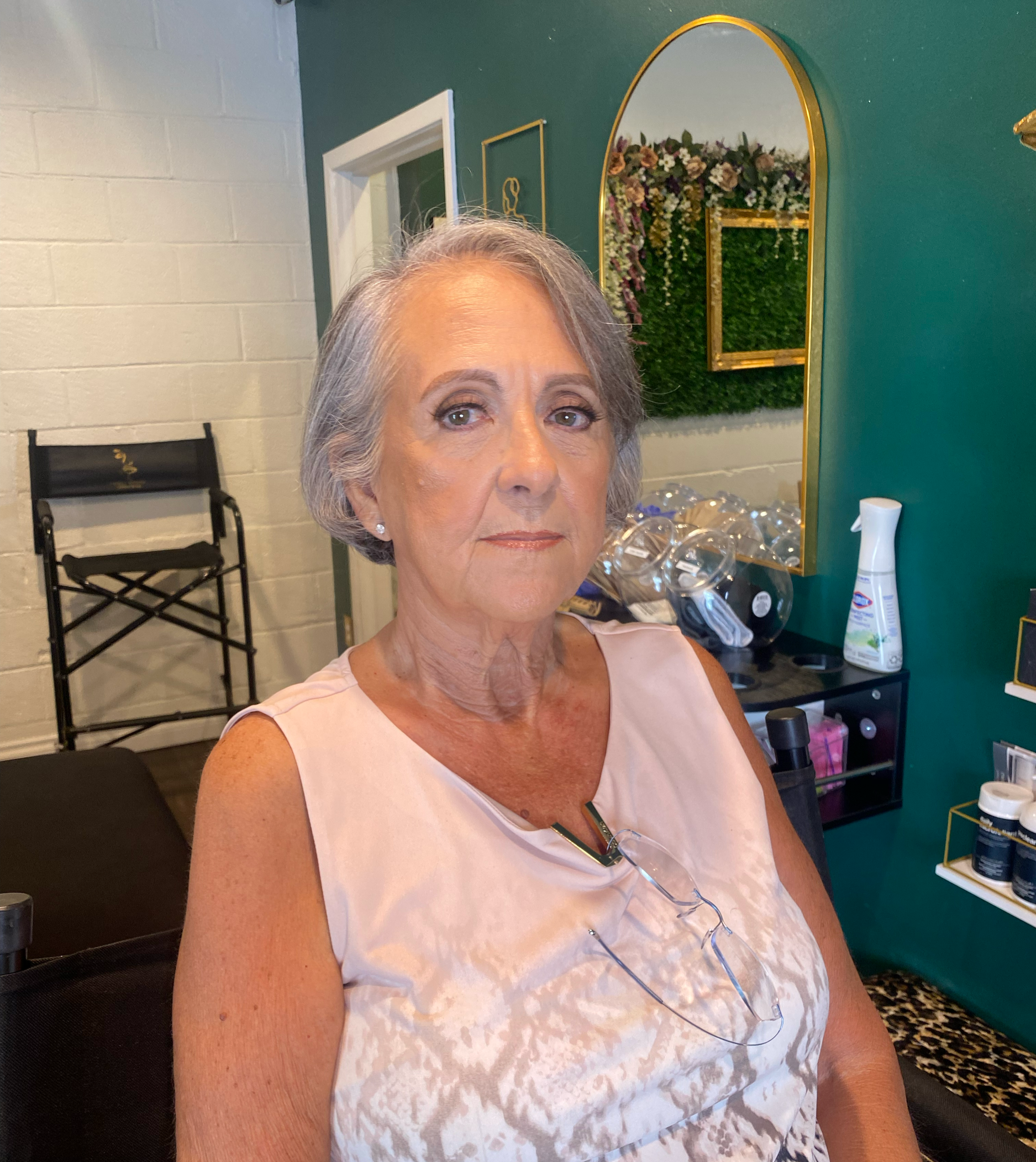 A woman with light gray hair and green eyes, wearing a sleeveless pale pink dress, is sitting in a chair in a room with green and white walls. There is a mirror with a gold frame on the green wall behind her, reflecting a floral arrangement and greenery. On the table beside her, there is a white device, and a black chair is visible in the background.