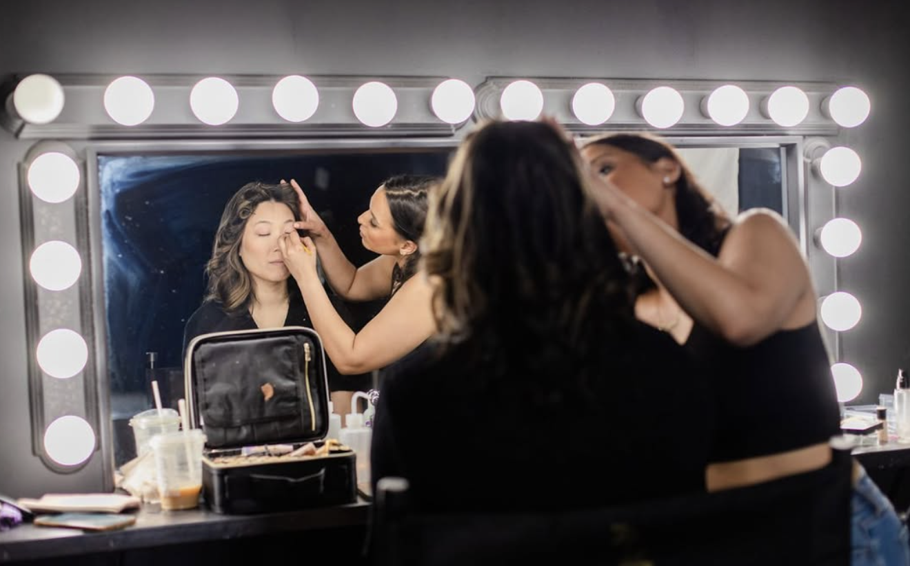 A makeup artist applying makeup on a woman in front of a mirror with bright vanity lights, with makeup supplies and drinks on the table.
