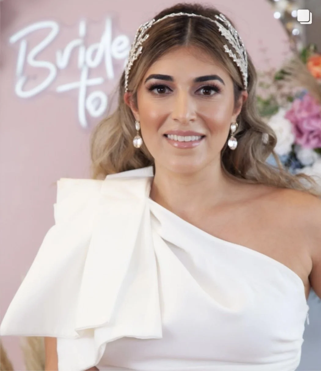 A woman with styled hair, wearing pearl earrings and a white dress with a large bow on the shoulder. She is smiling at the camera, with a pink background and flowers behind her. The sign in the background says 'Bride to be.'