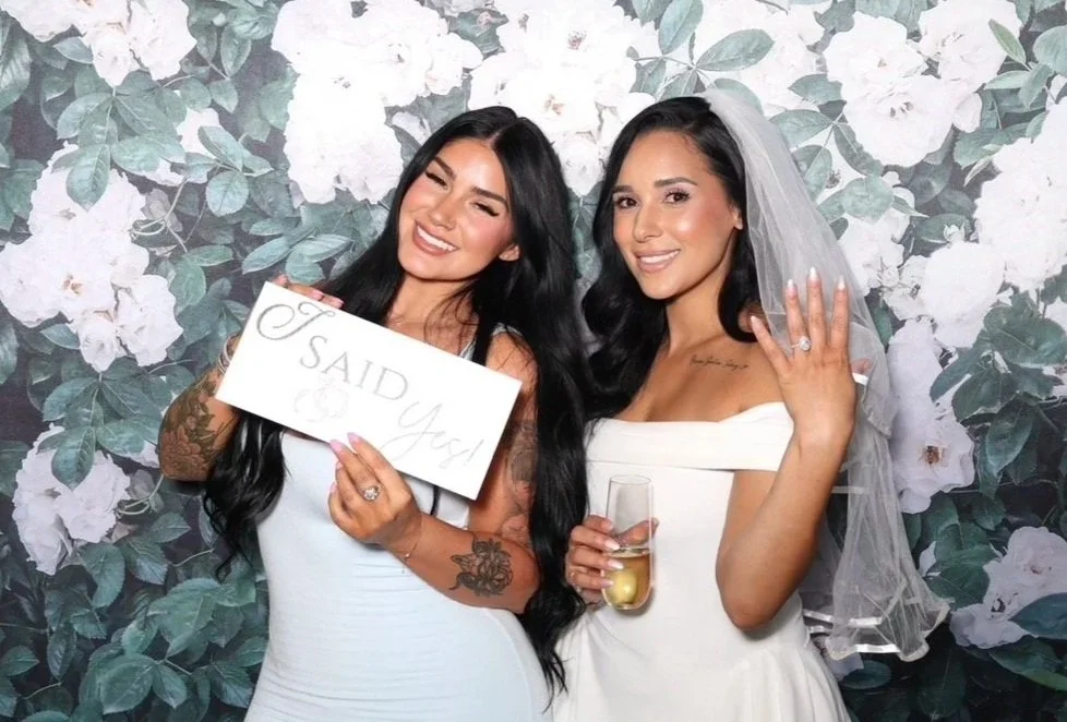 Two women, one in a white dress and veil, and the other in a white dress, at a wedding. The woman in white is showing her wedding ring with her left hand. The other woman is holding a sign that says "I Said Yes!" Both women are smiling and standing in front of a floral backdrop.