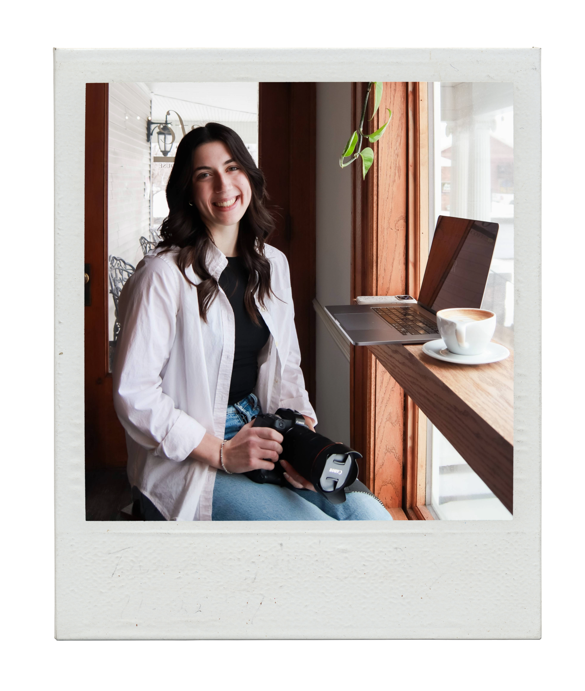 Young woman with dark hair smiling, sitting by a window in a cafe, holding a camera, with a laptop and a cup of coffee on the windowsill.