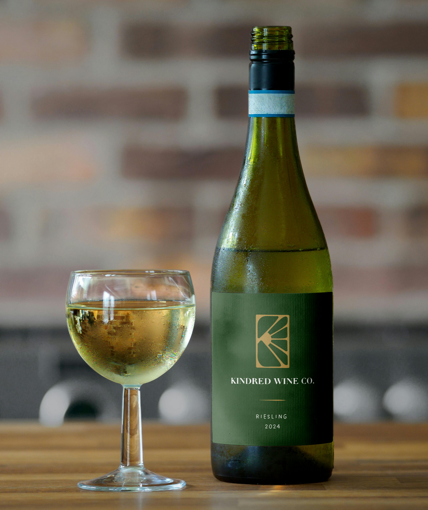 A bottle of white wine labeled 'Kindred Wine Co. Riesling 2024' and a glass of white wine on a wooden surface with a blurred brick wall background.