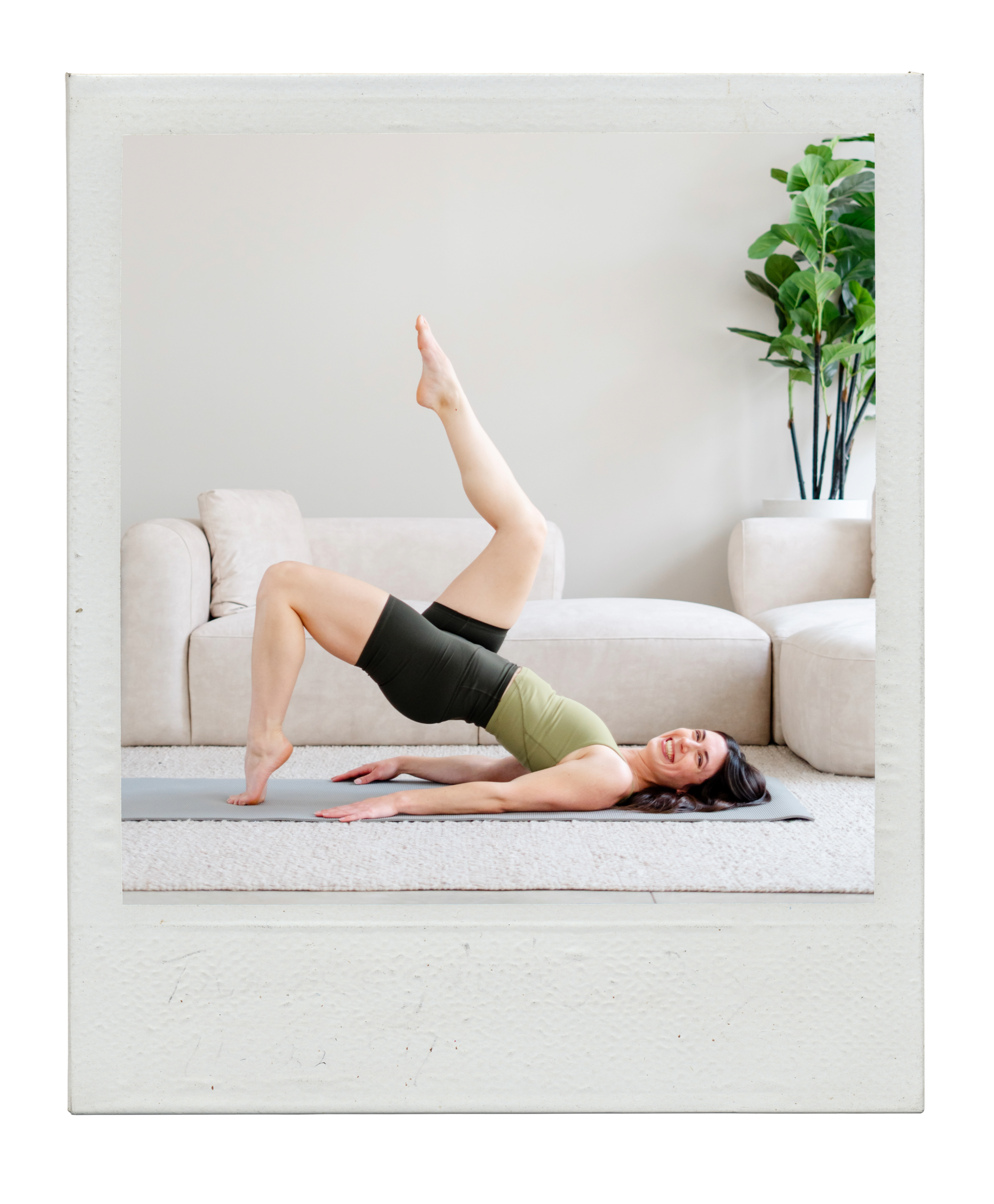 A woman practicing yoga in a living room, lying on her back with one leg raised vertically, smiling, wearing black shorts and a green top, with a white sofa and potted plant in the background.