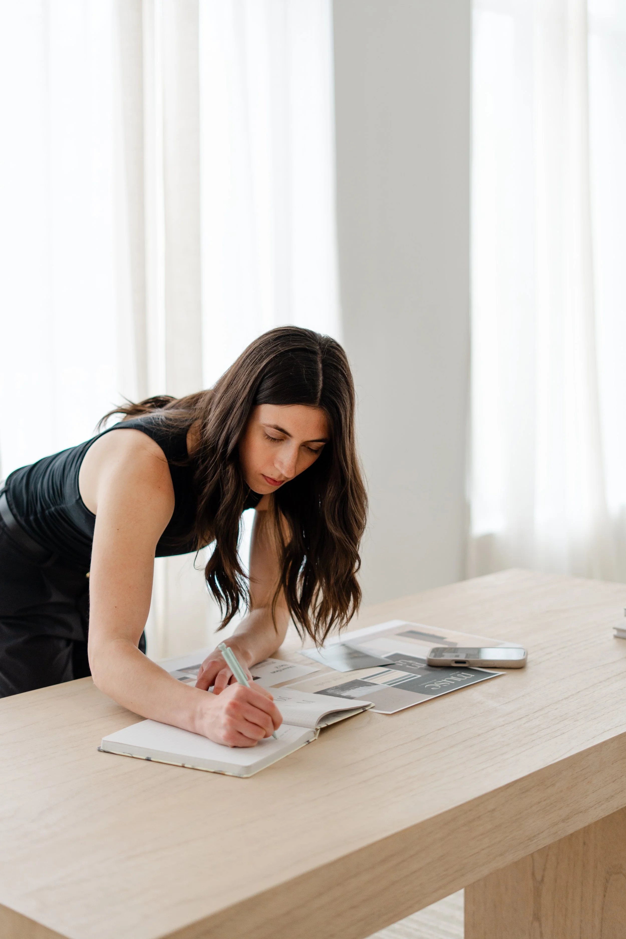 A woman with long, dark hair is leaning over a wooden table, writing in a notebook. There are magazines and a smartphone on the table in front of her, and she is wearing a black sleeveless top. Bright natural light is coming through the windows with white curtains behind her.