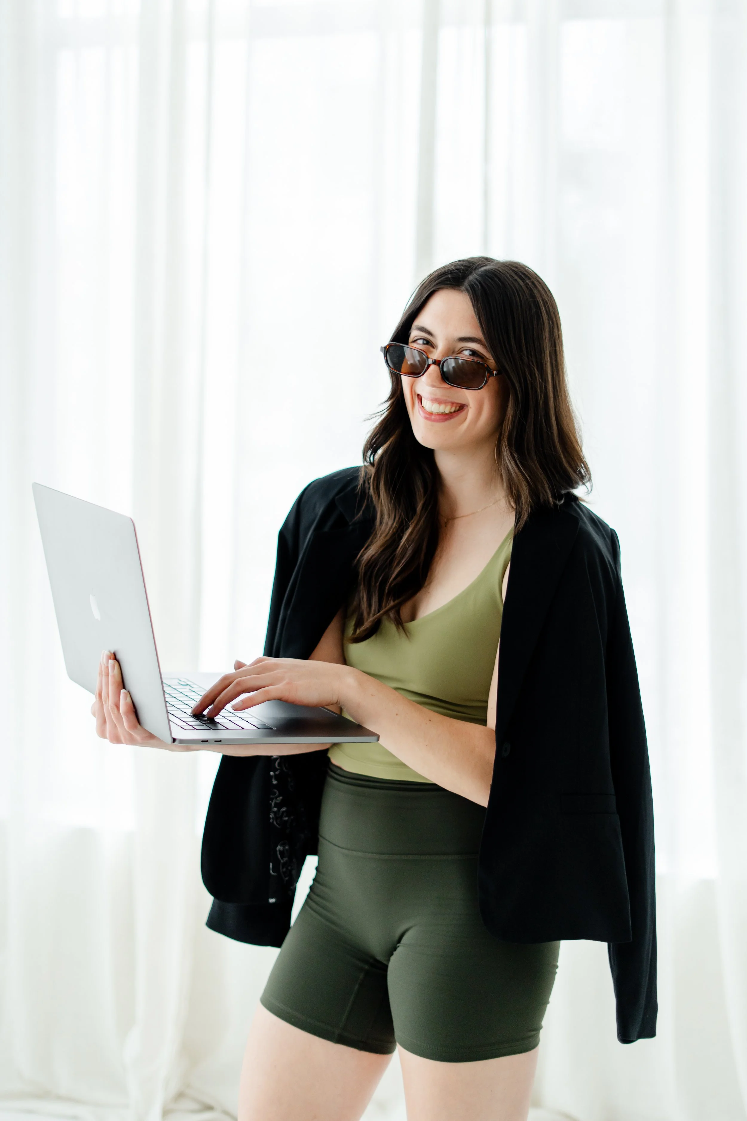 Young woman with dark hair wearing sunglasses, a green top, a black blazer, and gray shorts holding a silver laptop indoors.